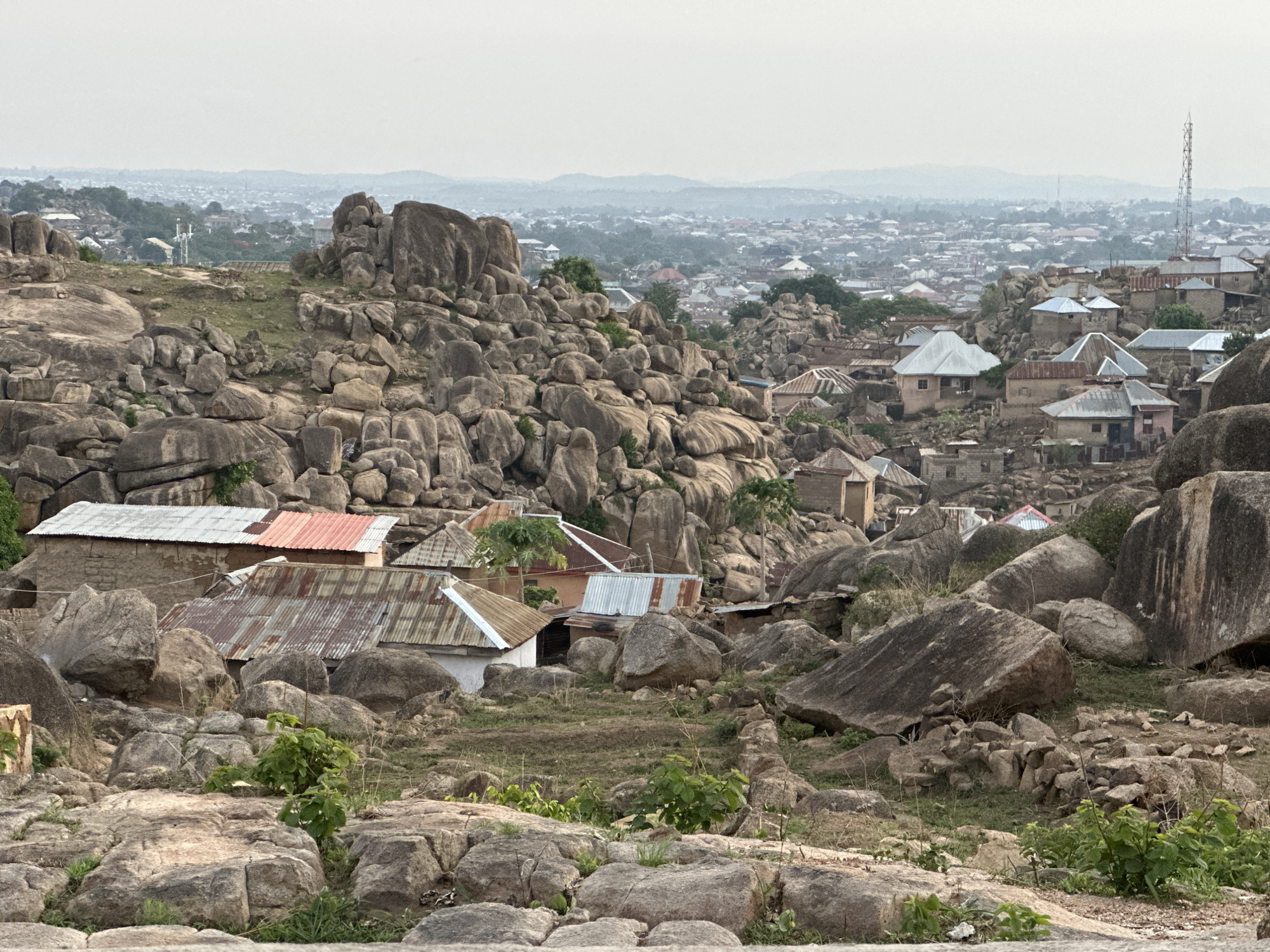 The image shows a view of a section of the city of Jos, Nigeria, known for its rocky landscape. The foreground is filled with large, grey boulders and sparse vegetation. Several houses with corrugated metal roofs are scattered among the rocks. In the background, a dense urban area extends towards the horizon, with a communication tower visible on the right side of the frame. The sky is overcast, giving the scene a muted tone. No people are visible.