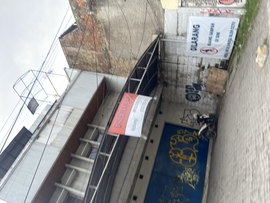 The image shows a narrow alleyway in Margahayu, Indonesia (latitude: -6.965, longitude: 107.577). The alley is lined with brick walls and buildings on both sides, with a metal awning overhead. A blue metal gate with graffiti painted on it can be seen in the background. There is a motorbike parked in the alley, facing away from the camera. On the right wall, there is a sign that reads "DILARANG BUANG SAMPAH DI SINI" in white text on a blue background. The sign is attached to a brick wall, and the wall is covered in a layer of dust and dirt. There are several power lines running across the alleyway.