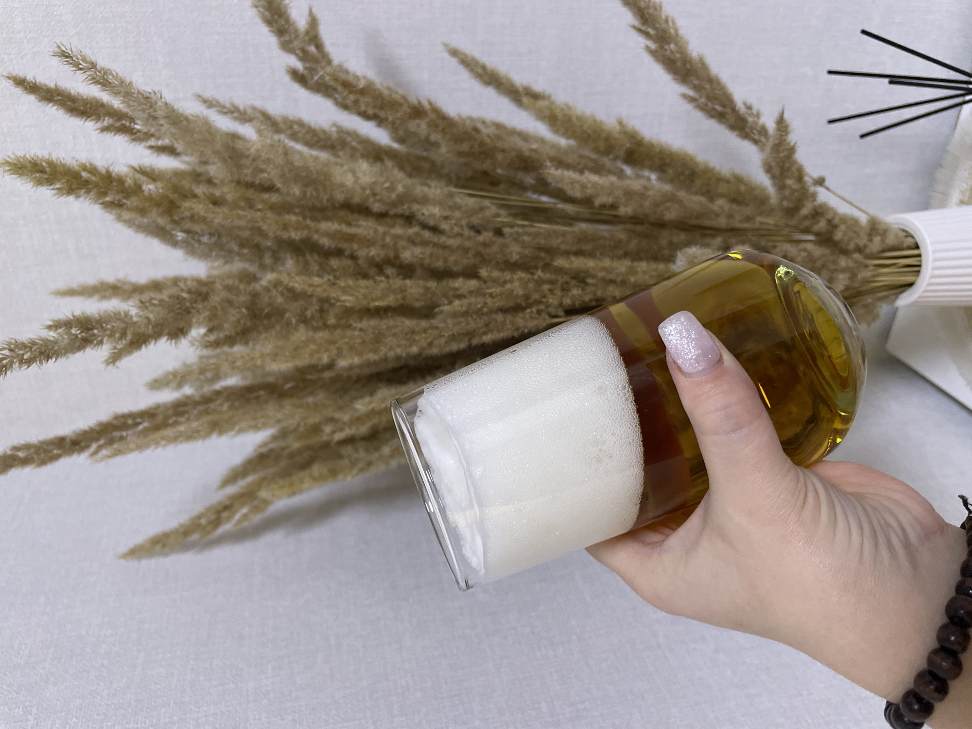A human right hand, with light skin tone and fingernails painted with a light pink base and white glitter tips, holds a clear, slightly tapered glass filled with a golden-amber liquid and a substantial layer of dense white foam on top. A dark brown beaded bracelet adorns the wrist. The background features a large bouquet of dried, feathery pampas grass (or similar ornamental grass) in a white, vertically ribbed ceramic vase, positioned on a light-colored surface. To the far right, partially visible, are several dark, slender diffuser reeds. The overall background consists of a light, textured wall. The scene is brightly lit, indicating an indoor environment.