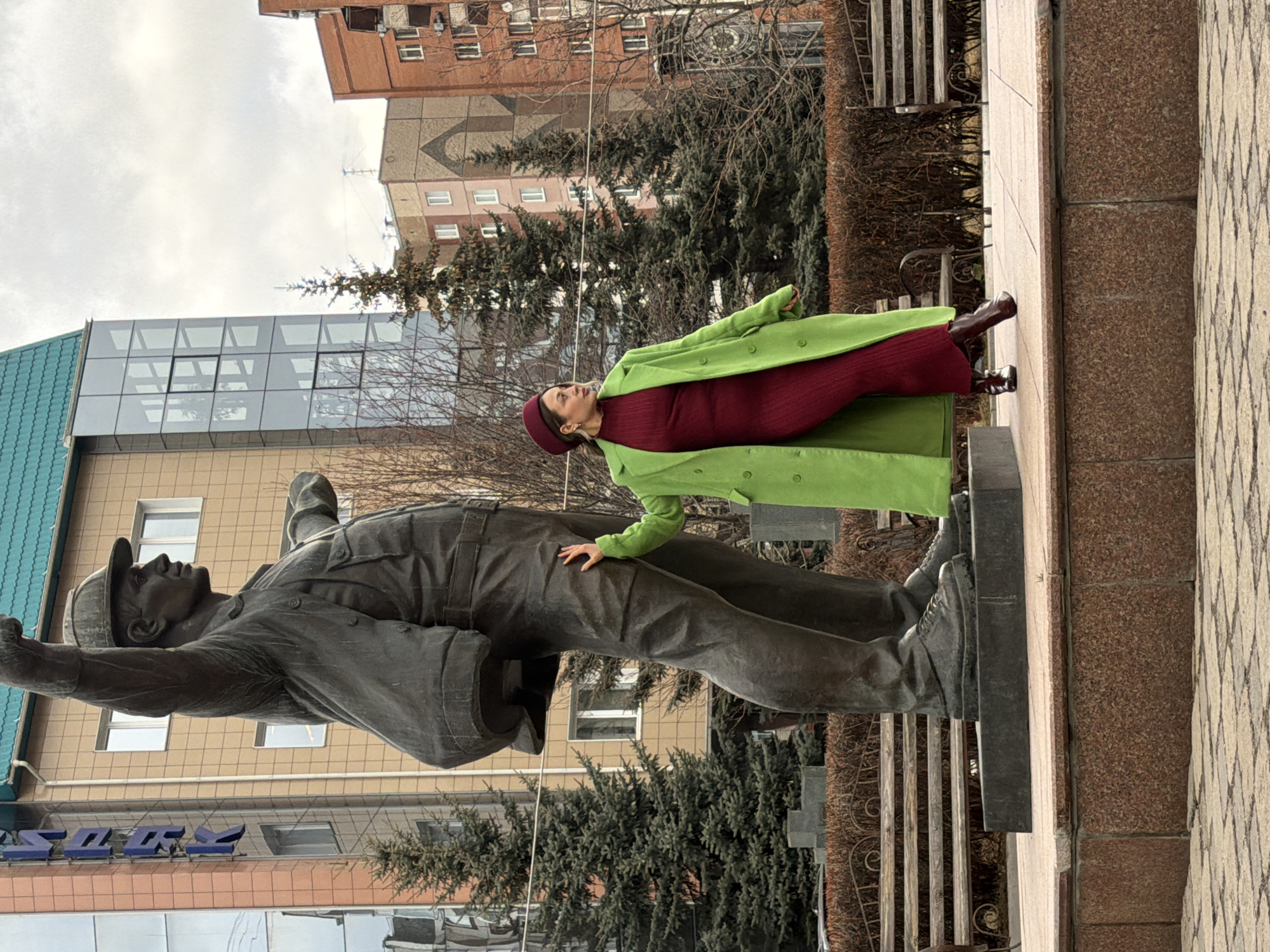 A woman in a green coat and burgundy dress poses with a bronze statue of a worker in what appears to be Krasnoyarsk, Russia. The woman, wearing a beret of the same color as her dress, leans against the statue, one hand resting on its shoulder. The statue depicts a worker raising one arm and fist in the air. Buildings can be seen in the background, including a modern glass structure and a brick building. The weather appears overcast. The statue is located on a brick base next to a paved area.