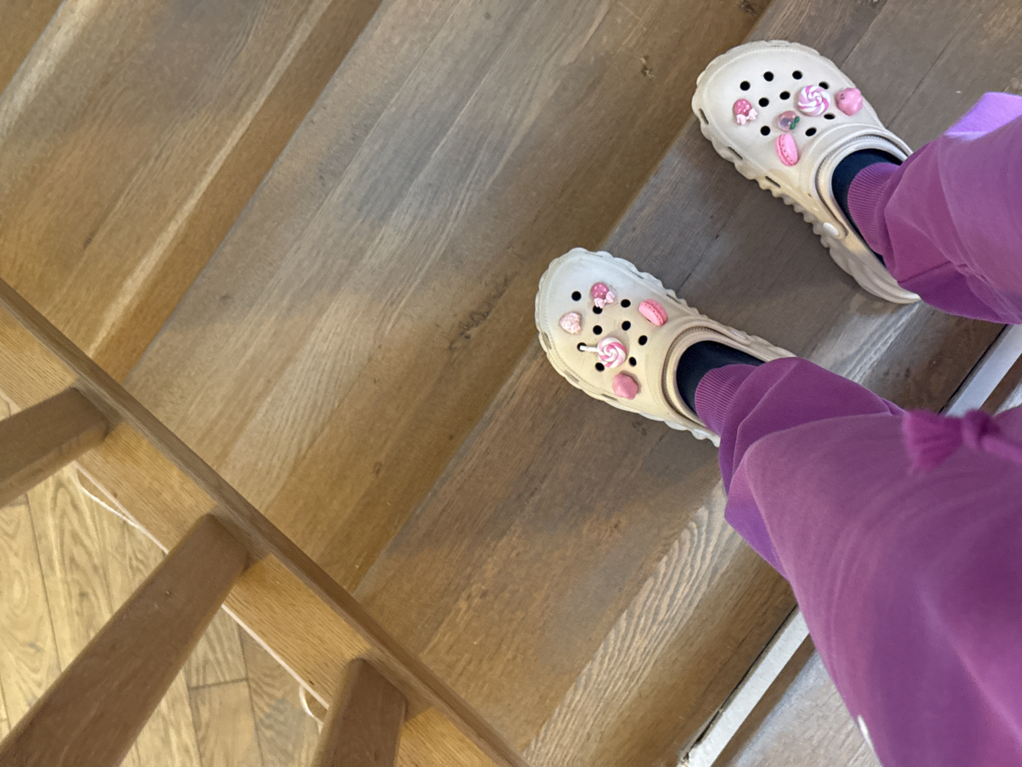 The image depicts a person standing on a wooden staircase. The person is wearing light-colored Crocs adorned with candy-themed charms, paired with dark socks and purple sweatpants. The wooden stairs appear to be made of oak. The location is unknown and cannot be determined from the image.