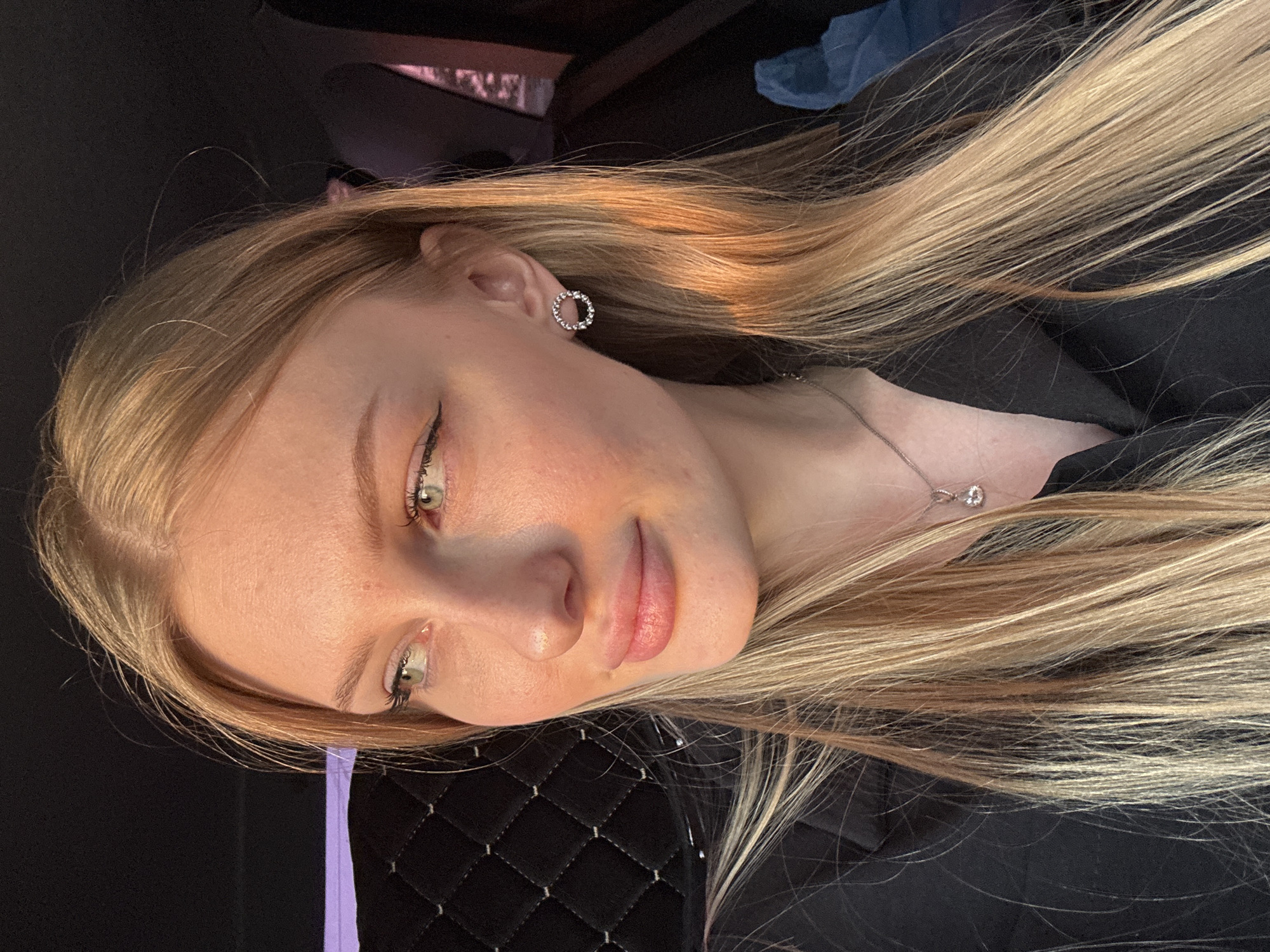 A young woman with light blond hair, fair skin, and green eyes, wearing a silver necklace and hoop earrings, is the central figure in this photo. She is wearing eye makeup with eyeliner. The background includes dark upholstery and a hint of what appears to be an interior space with lighting, possibly a vehicle, which suggests she may be in a car in or near Kondratovo, Russia.