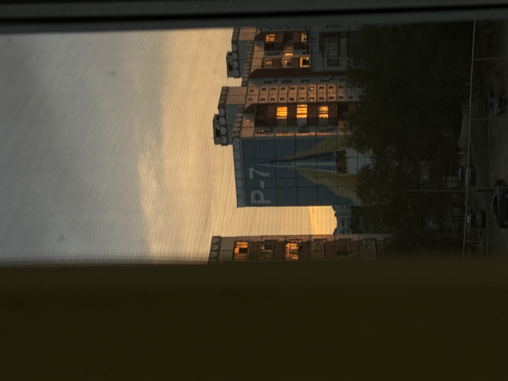 A vertical, eye-level, full shot captures an exterior scene in Baikonur, Kazakhstan, viewed through a window with a screen. The image is dominated by the warm hues of a sunset sky, with soft oranges and yellows blending into a muted gray. In the foreground, the left side of the frame is obscured by a dark, out-of-focus object, likely a curtain or wall.
The middle ground features several multi-story apartment buildings. The closest building on the left is tall and narrow, with many windows, some of which reflect the warm sunset light. To its right is a wider, larger building adorned with a large blue mural featuring a white rocket and the letters "P-7" in white. This building also has numerous windows reflecting the sunset, creating a striking contrast with its darker facade. To the right of this, another apartment building, slightly further away, also shows windows illuminated by the setting sun.
The lower portion of the image depicts a street scene. Trees with dark foliage are visible in front of the apartment buildings. Several cars are parked or driving on the street, and a low fence runs along the bottom of the frame. The overall impression is a view from an apartment window overlooking a residential area during sunset, with the iconic imagery of the "P-7" rocket mural on one of the buildings hinting at the location's connection to space exploration, a key characteristic of Baikonur.