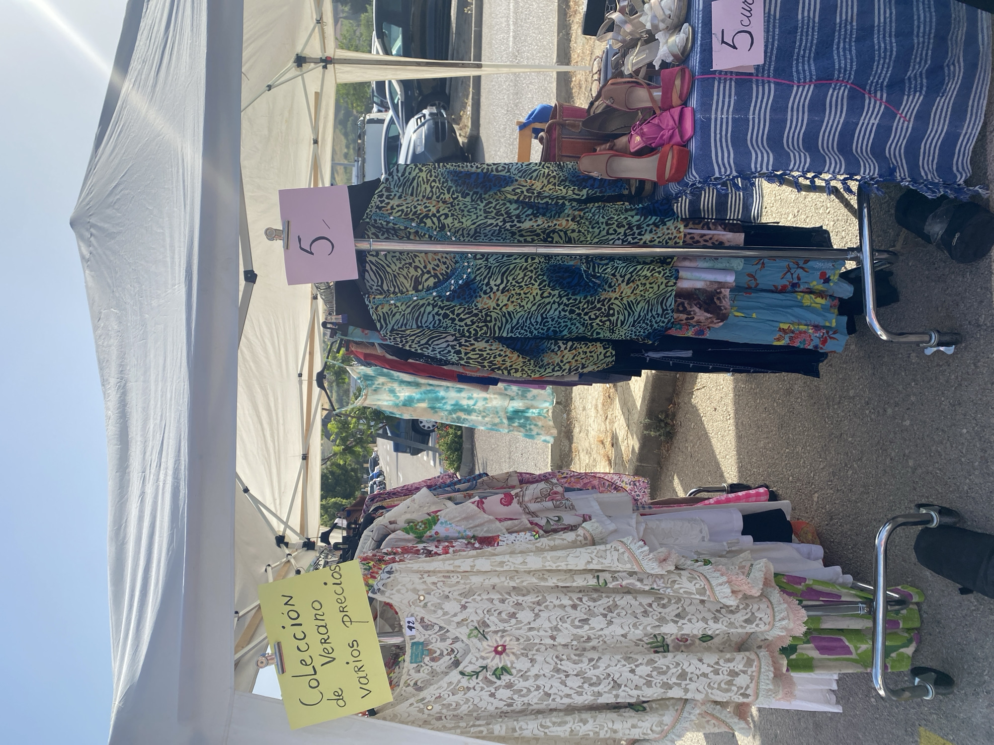 This is a medium shot, vertically oriented photograph of an outdoor market stall. The stall is covered by a white canopy, with part of a blue and white striped cloth draped over it on the right. Racks of clothing are displayed for sale. In the foreground, a white lace top with floral embellishments is visible. Behind it, various other garments, including patterned shirts and dresses, are hung or folded. On the left, a yellow sign reads "Colección de Verano varios precios" in black lettering. A pink sign with a black "5" is attached to the clothing rack on the right.
The scene appears to be a street or sidewalk market. The background shows a paved road and some greenery, with cars parked further back. The lighting suggests it is a sunny day, likely morning or afternoon, with bright sunlight casting shadows. The overall impression is of a casual, outdoor shopping experience, possibly a flea market or second-hand clothing sale. No people are clearly visible in the frame, but the presence of the stall and merchandise indicates a commercial activity. The context of Manilva, Spain suggests a Mediterranean setting.