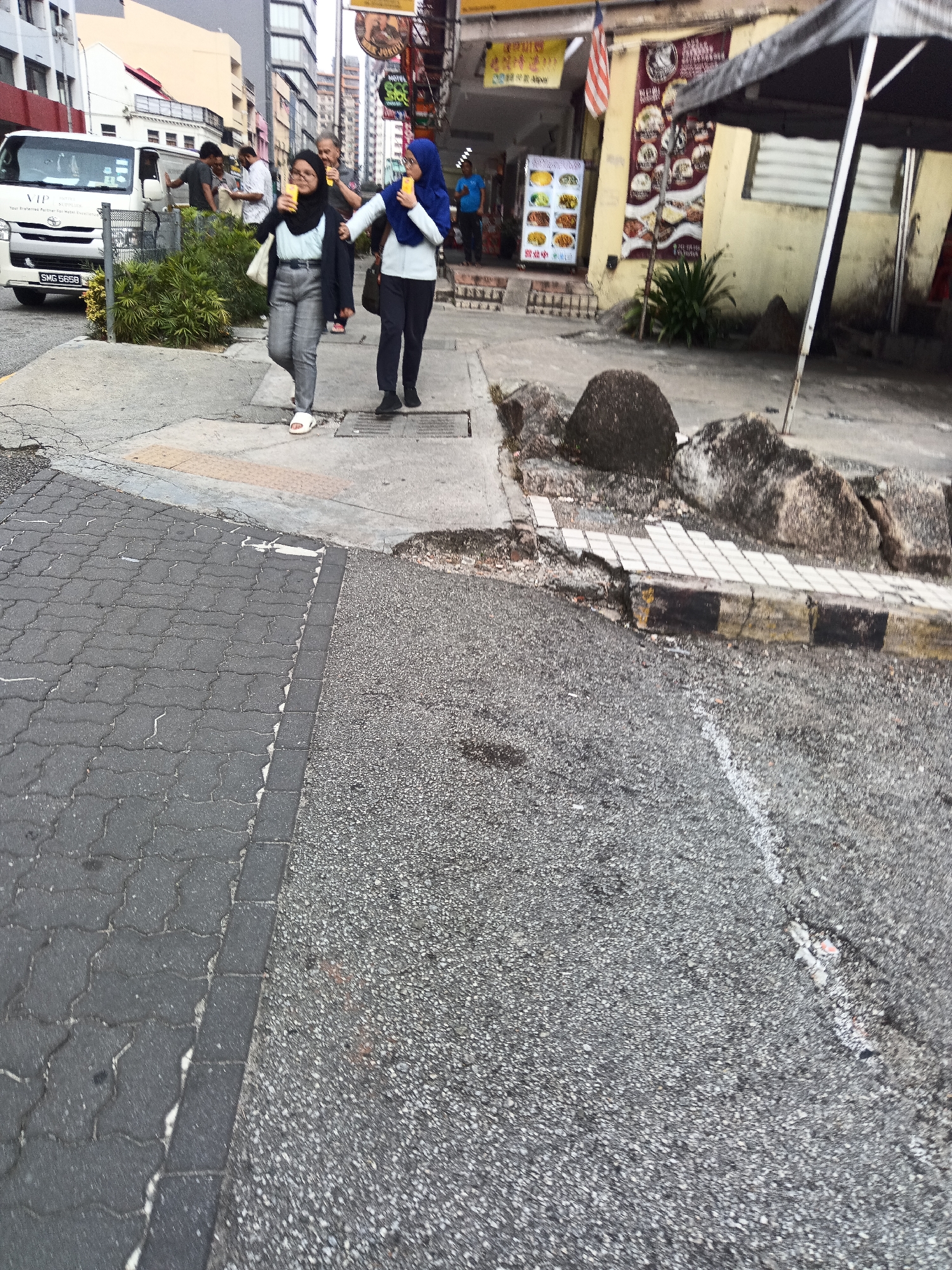 In Kuala Lumpur, Malaysia, two women in headscarves walk side-by-side on a sidewalk next to a busy street. The woman on the left wears a dark headscarf and a black blazer over a white top and grey pants. She carries a white tote bag. The woman on the right wears a blue headscarf and a light blue top with dark pants. Both women are holding yellow cups. In the background, a white van with "VIP" branding is parked near a row of buildings with storefronts. Several other people are visible on the sidewalk and near the van, suggesting a bustling urban environment. The street is paved with cobblestones and asphalt.