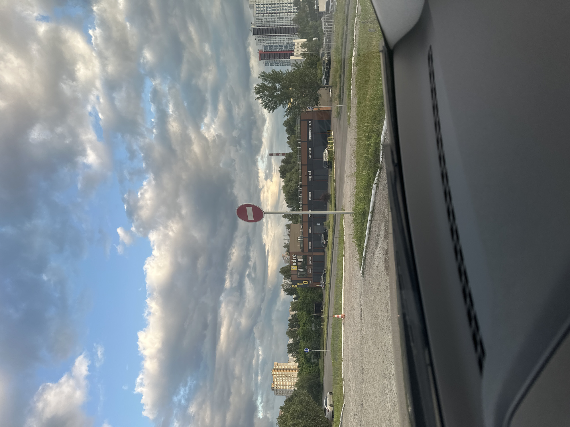 The image depicts a street scene in Kyiv, Ukraine, under a cloudy sky. In the foreground, the dashboard of a car is visible, indicating the photo was taken from inside a vehicle. The road ahead is paved, with grass and a white line marking the edge. Further into the scene, a single red and white traffic cone is placed on the pavement. To the right, a white signpost with a red circle and a horizontal white bar, indicating a prohibition, stands tall.
Beyond the road, a low-rise commercial building is visible with the text "adre STO" on its facade, alongside words such as "РЕМОНТ" (repair), "ХОДОВОЙ" (undercarriage), "ЗАПЧАСТИ" (parts), and "ШИНОМОНТАЖ" (tire fitting). Several cars are parked in front of the building and on the side of the road. A few trees are planted along the building's frontage and behind it.
In the mid-ground, tall residential buildings are visible on the right side of the frame. On the left, a taller, lighter-colored apartment building stands amongst trees. A red and white striped chimney is also visible in the distance. The overall atmosphere is overcast, suggesting it might be late afternoon or early evening.