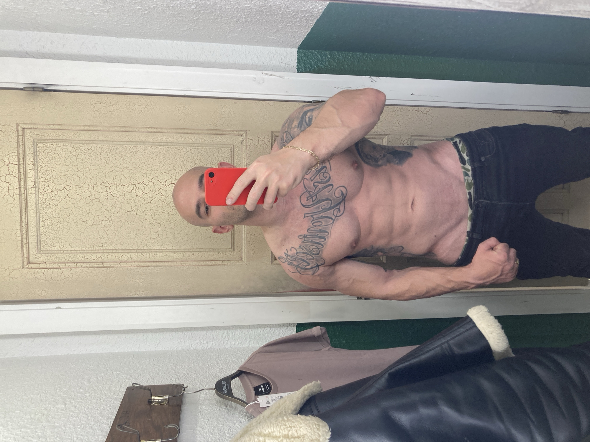 The image is a mirror selfie depicting a bald, muscular adult male, shirtless and tattooed extensively across his chest and left arm. He holds a red smartphone in his right hand, obscuring his face, and wears a gold-colored chain bracelet on his right wrist. He is dressed in dark jeans, with a camouflage-patterned waistband visible above the denim. His left hand is clenched into a fist. The setting is an interior room, likely a fitting room. The wall on the viewer's left is white, while the wall on the viewer's right is split horizontally, white on the upper section and dark green on the lower. Behind the subject, an old wooden door with cracked, light-colored paint is visible. A wooden coat rack with brass-colored hooks is mounted on the left wall, from which hangs a light pink short-sleeved t-shirt on a "PULL&BEAR" branded hanger with a visible price tag, along with a dark leather-like jacket featuring cream shearling trim. The presence of a "PULL&BEAR" branded item indicates the location is a fitting room within a Pull&Bear retail store in Sofia, Bulgaria.