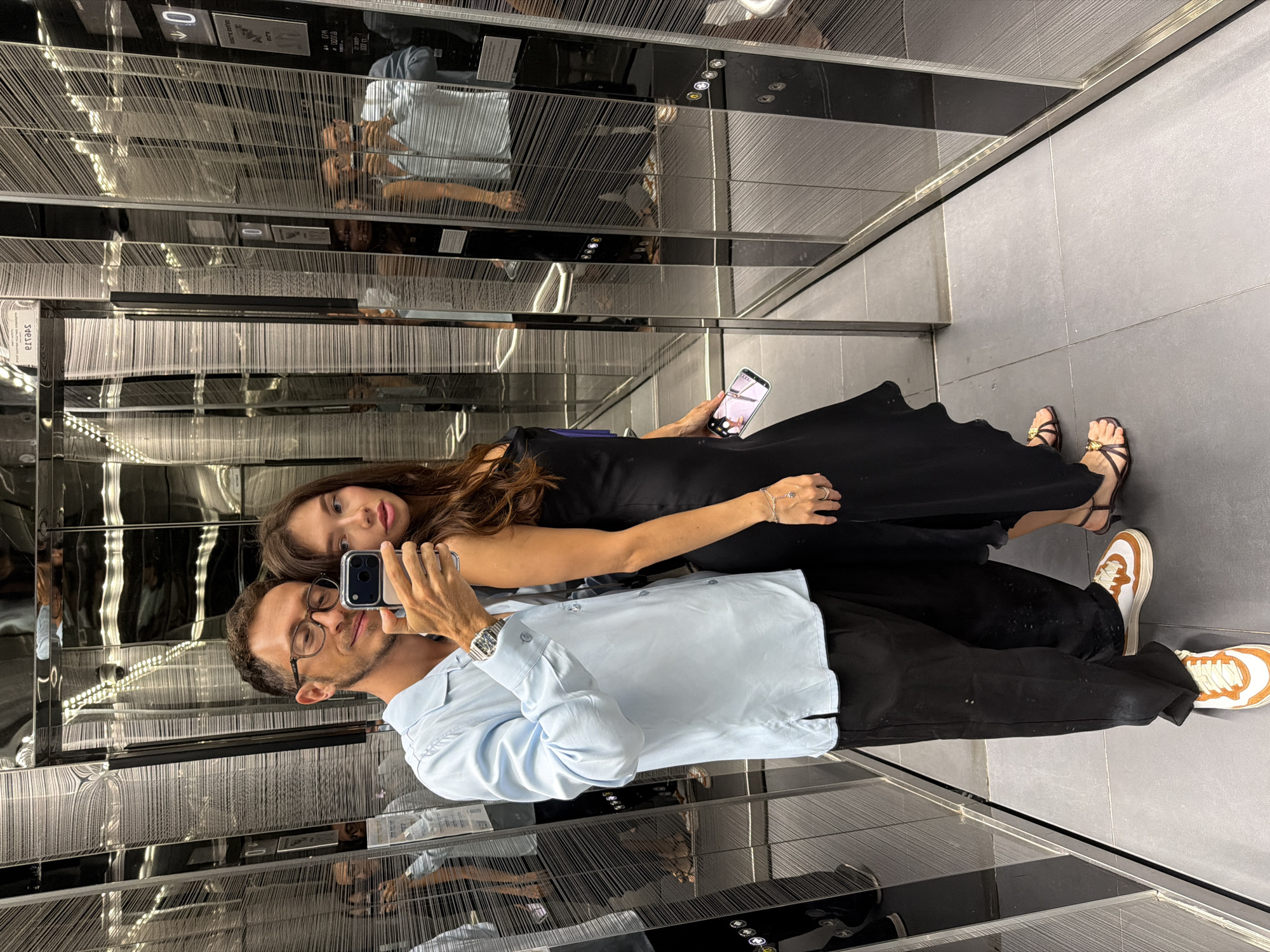 The image was taken in an elevator in Tel Aviv, Israel. A couple is posing for a mirror selfie. The man is wearing glasses, a light blue shirt, and black pants. The woman is wearing a black dress and sandals. They are both holding their phones, and the woman is also holding another phone in her left hand. The elevator walls are reflective, showing multiple reflections of the couple and the elevator interior. The floor is tiled in a neutral gray color.