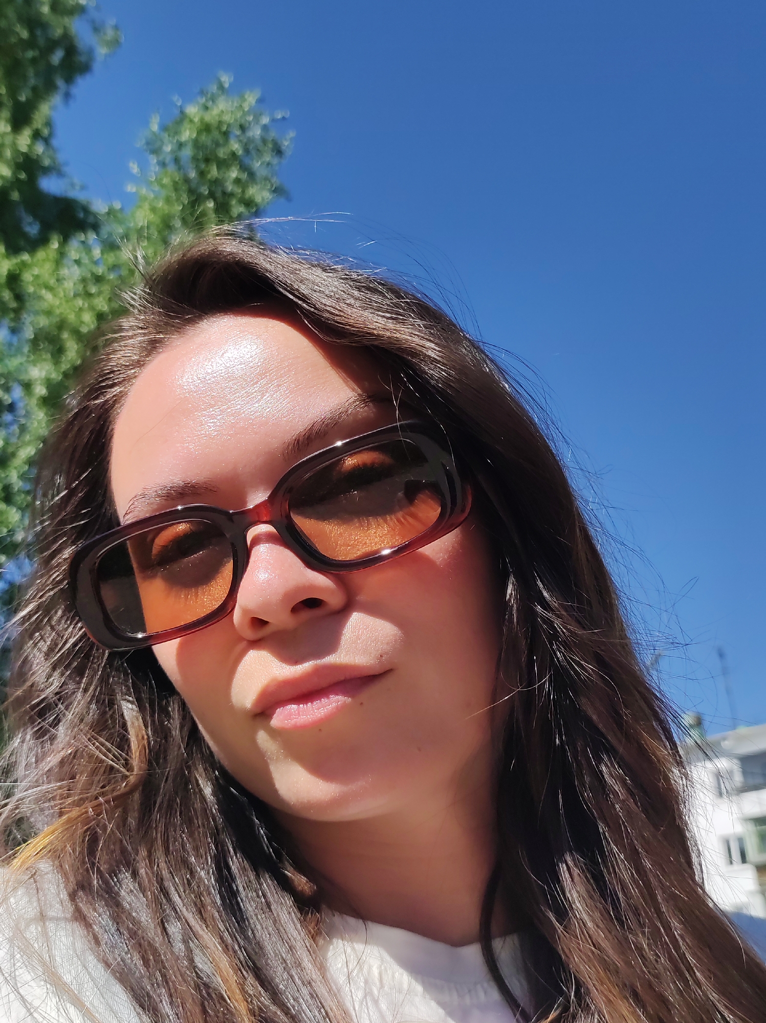 A woman with long brown hair is pictured in an upward-facing selfie. She is wearing brown-framed sunglasses with amber-tinted lenses. The background includes a clear blue sky and the green foliage of trees, suggesting an outdoor setting. In the lower right of the frame, a portion of a white building with visible windows is present. The image was most likely taken in a sunny outdoor location. The city of Chaikovsky is not definitively verifiable from this image.