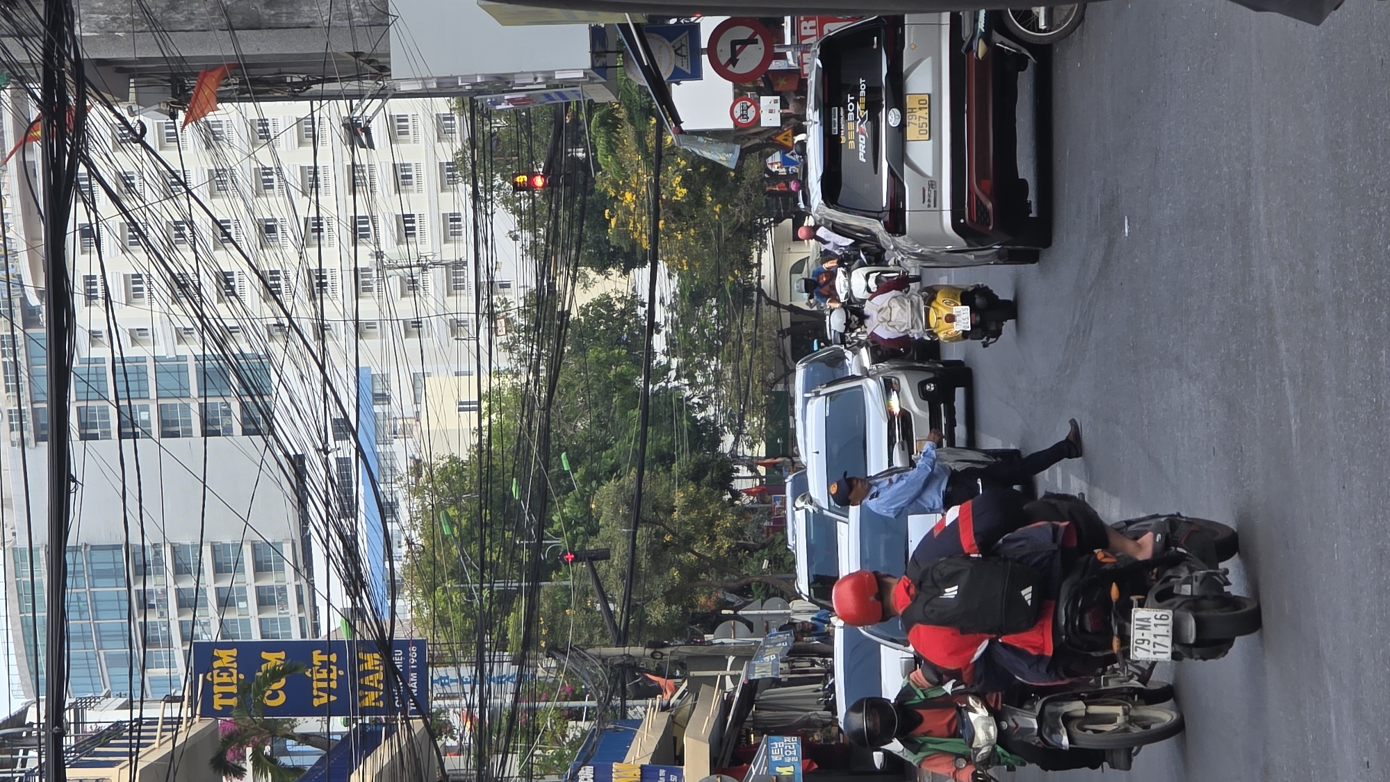 This image captures a bustling street scene in an urban area, likely Nha Trang Ward, Vietnam, during daylight hours. The perspective is from the side of the road, looking across multiple lanes of traffic and towards buildings and greenery in the distance.
The foreground is dominated by a dense network of overhead electrical and communication wires crisscrossing the sky, a common sight in many Vietnamese cities. Below these wires, a street-level view shows a mixture of vehicles, predominantly motorcycles and a few vans/cars, stopped or moving slowly in what appears to be heavy traffic, possibly at a red light given the red signal visible on a distant traffic light.
People are visible on motorcycles, wearing helmets and everyday attire. One motorcyclist in the immediate foreground wears a red helmet and jacket with a black backpack, while another behind them has an orange/green vest. A man in a blue shirt is seen standing near a white van.
In the midground, beyond the vehicles, there are lush green trees and some trees with yellow blossoms, providing a natural contrast to the concrete environment. Several multi-story buildings are visible in the background, including a large white building with numerous windows and another building with blue-tinted windows.
Visible text includes:
* A blue sign on the left with "TIỆM CƠM VIỆT NAM TỪ NĂM 1900" which translates to "VIETNAMESE RICE RESTAURANT ESTABLISHED IN 1900."
* A motorcycle license plate "79-NA 171.16".
* On the back of a white van, "BEEBOT PRO" and "BOT" are visible, along with the license plate "79H 057.10".
* Several street signs are visible, including a "no left turn" sign and a "no entry" sign.
The overall impression is one of a vibrant, active urban environment with typical Southeast Asian street characteristics like dense wiring and a mix of motorbikes and cars.