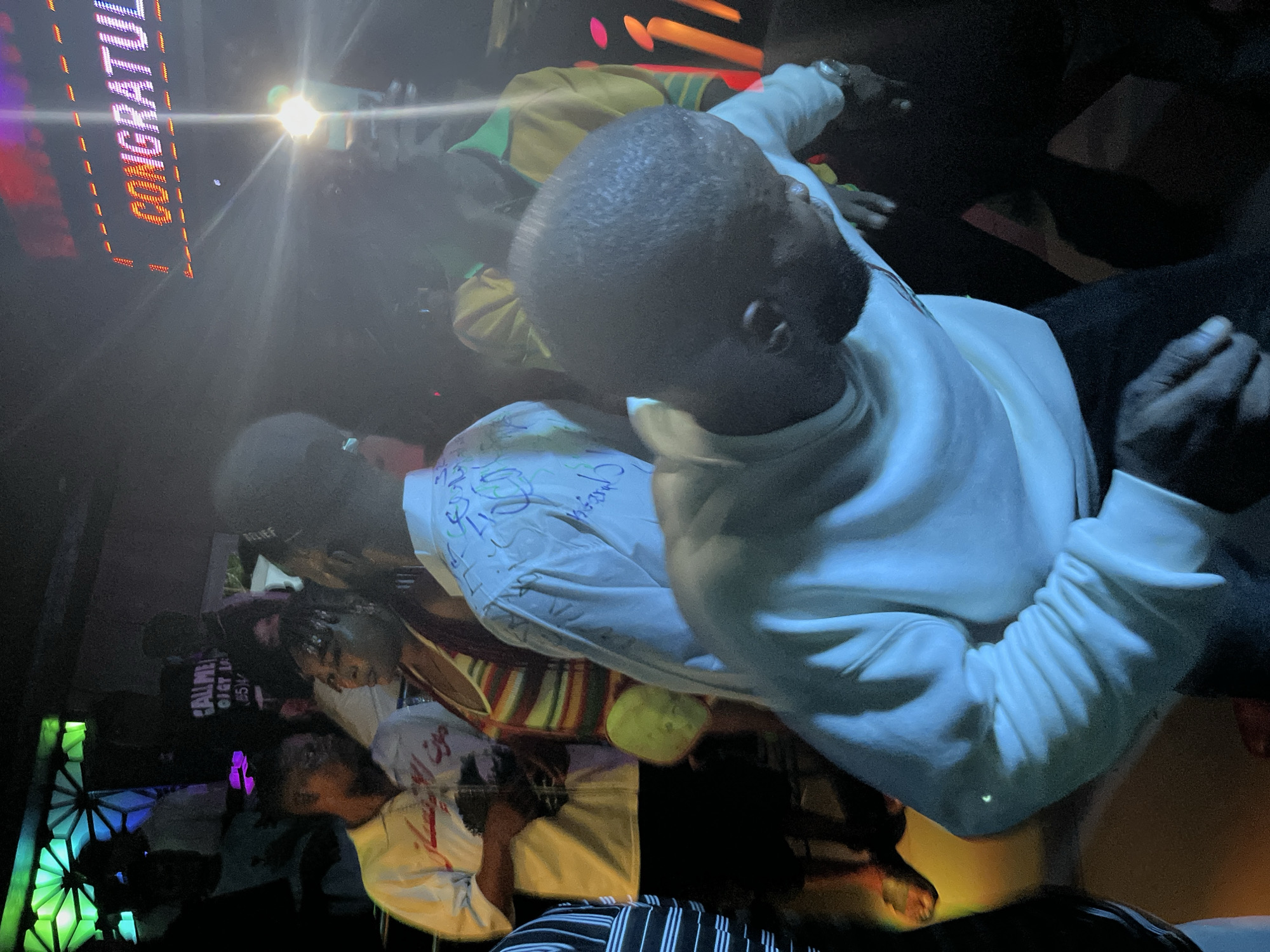 In an indoor nighttime setting, a crowd of people is gathered, illuminated by various lights. In the background, a brightly lit sign displays the word "CONGRATULAT." Several individuals are visible, some in the foreground and others further back. One person in the foreground wears a white long-sleeved shirt with blue and green writing on it and appears to be dancing or moving. Another person in the midground is wearing a white shirt with writing on it and is facing the right side of the frame. To their left, a person in a striped top is visible. The environment is dimly lit, with splashes of color from the lighting and the sign. The overall atmosphere suggests a celebratory event or party.
