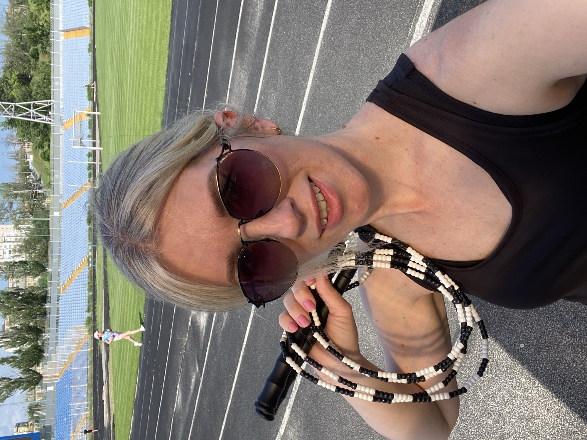A woman with blond hair and sunglasses holds a jump rope with black and white beads at an outdoor track and field stadium. She is wearing a black tank top. A person is running on the track in the background. The stadium has blue seats. The location is in the city of Unknown, Unknown.