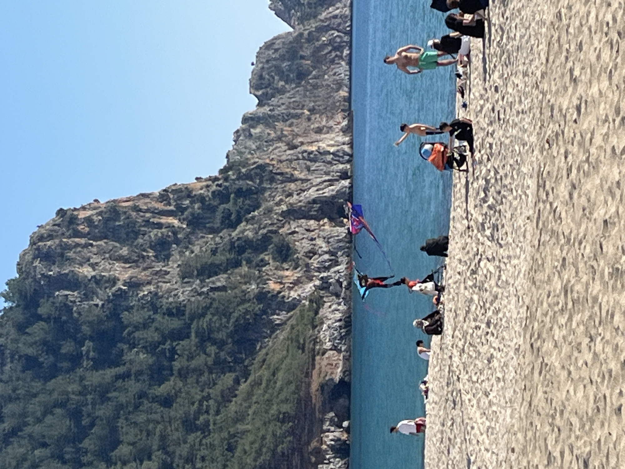 The image depicts Cleopatra Beach in Alanya, Turkey. The foreground consists of light-colored sand with numerous undulations and visible footprints. The midground features clear, calm turquoise water of the Mediterranean Sea. The background comprises a large, steep landmass: densely tree-covered hills on the left transition into rugged, exposed rocky cliffs and formations on the right, all beneath a clear blue sky.
Approximately 17 individuals are visible within the scene. In the water, two individuals are engaged in watersports, each connected to a large, steerable kite; one kite is blue and black, the other pink and purple. On the sandy beach, a male in a white shirt and red shorts stands near the water on the far left. Several individuals are seated or reclined on the sand to his right. Further right, a male in green swim shorts stands at the water's edge. To his left, a boy in dark shorts stands in the water, raising his right arm. An orange and blue object, possibly a flotation device or small seat, rests on the sand near the water. Additional individuals are dispersed across the central and right portions of the beach, observed standing, sitting, or resting. One individual in black attire is seated facing left, and another in black and white attire is partially visible standing further right.