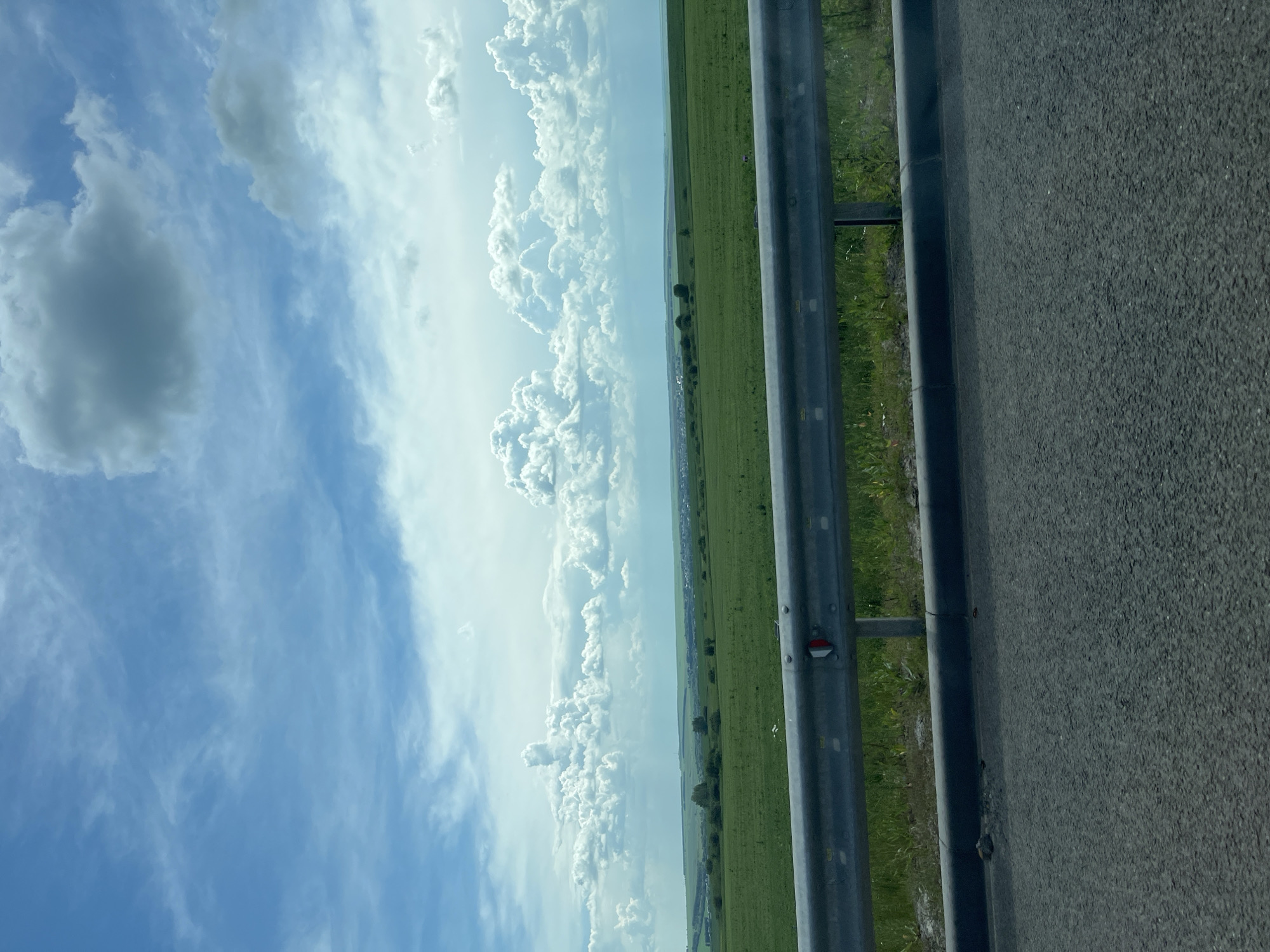 The image depicts a wide, green agricultural field stretching towards a distant horizon, under a partly cloudy blue sky. A metal guardrail runs horizontally across the lower portion of the frame, separating the road from the field. The road surface is visible in the foreground. The overall scene is rural, with no specific landmarks indicating the city of Kulikovka, Russia, beyond the provided context. There are no people or discernible actions taking place within the frame. The environment is characterized by open space and agricultural land.