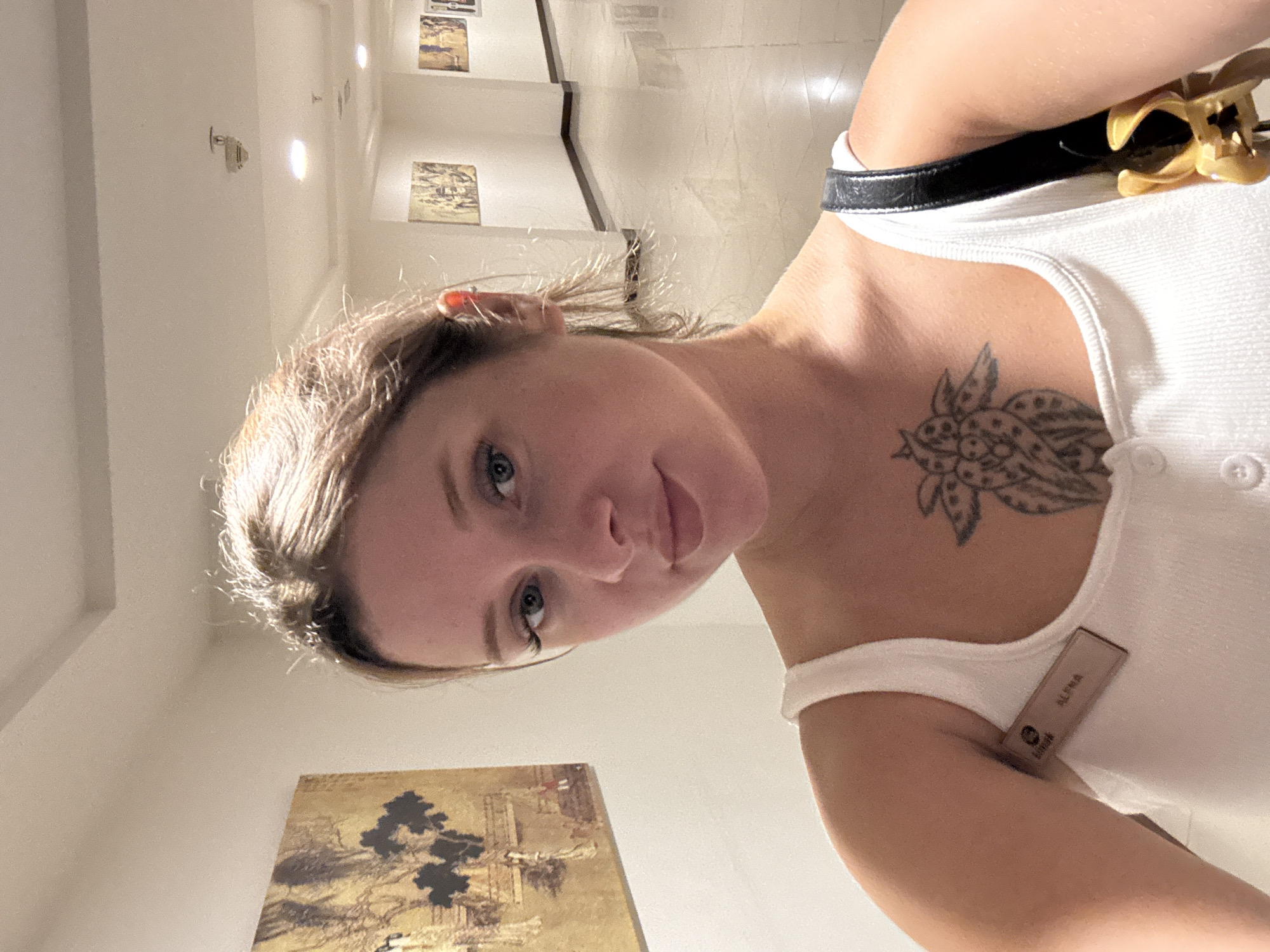 A selfie of a woman with blonde hair, blue eyes, and fair skin is taken in a well-lit hallway, possibly a hotel. She is wearing a white tank top with a name tag that reads "ALENA" and a tattoo of a flower on her chest. She is also carrying a black bag with a gold handle. The hallway features artwork on the walls and a tiled floor, suggesting an indoor, possibly public, location. No specific details of Aksu, Turkey, are visible in the image.