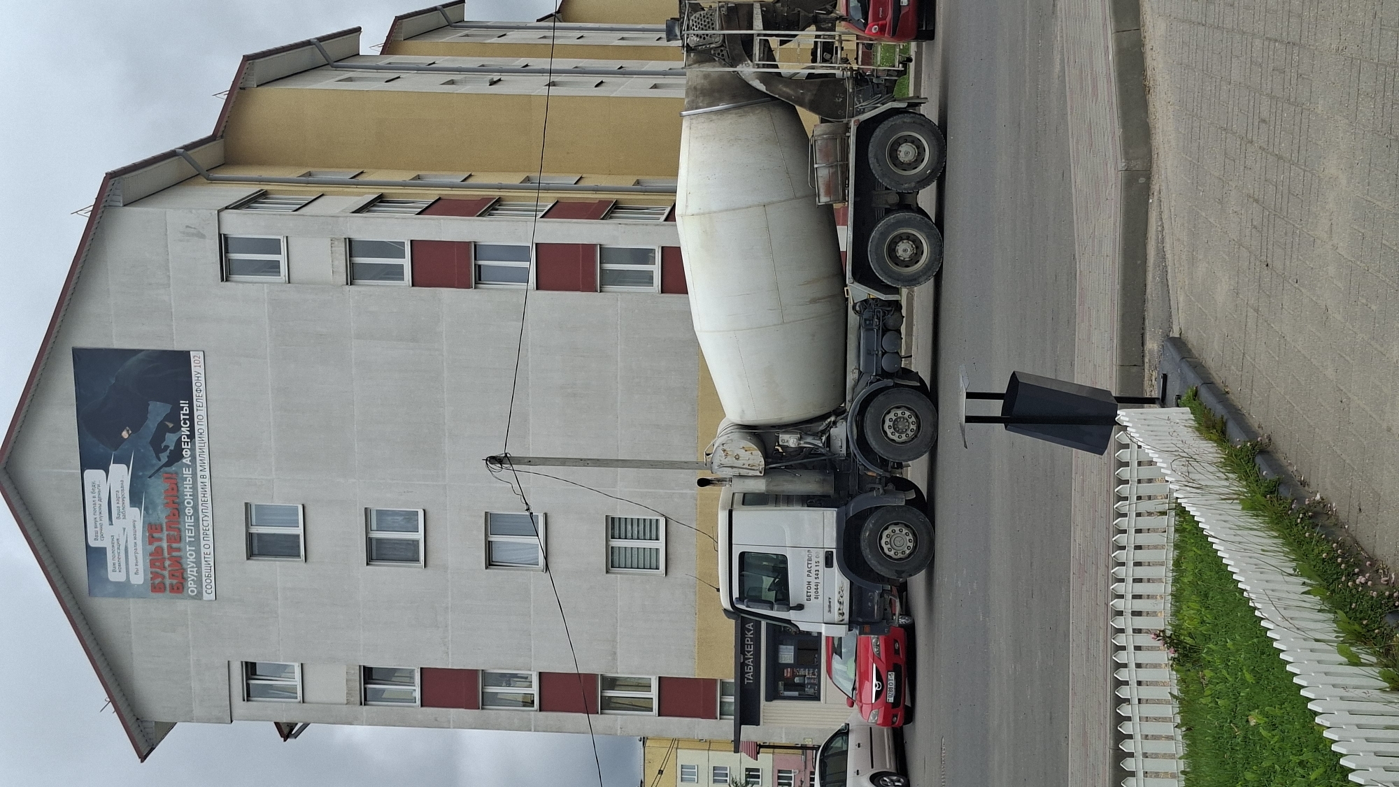 This is an outdoor, eye-level, full-shot photograph taken on a cloudy day in the city of Smolevichi, Belarus. The main subject is a white concrete mixer truck parked on a paved road in front of a multi-story apartment building. The building has a light gray facade with red trim around some of the windows and balconies. A large billboard is affixed to the top of the building, depicting a hooded figure holding a cell phone and warning about telephone scams, with text that translates to "BE CAREFUL! TELEPHONE SCAMMERS! REPORT CRIMES TO THE POLICE AT TELEPHONE 102."
To the left of the truck, a red sedan is parked, and further to the left, a portion of a white car is visible. A black trash can is positioned on a sidewalk to the right of the truck. In the foreground, a white picket fence encloses a patch of green grass with some small wildflowers. The pavement is a mix of asphalt and paving stones. The truck has text on its side that reads "БЕТОН.РАСТВОР" and a phone number.