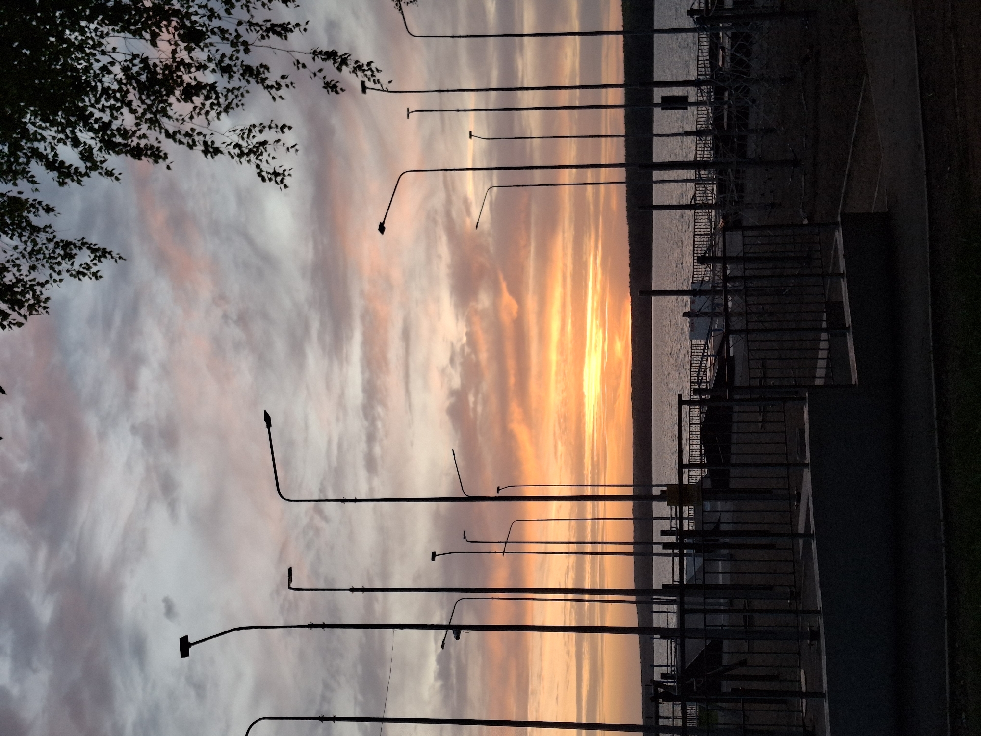 The image shows a sunset in Chaikovsky, Russia, taken from a waterfront area. The sky is filled with colorful clouds in shades of orange, pink, and gray. Silhouetted streetlights line the waterfront, with a metal railing separating the walkway from the water's edge. Dark trees appear in the upper left of the frame. The water reflects the colors of the sunset. A walkway and adjacent grass are seen on the right side of the image.