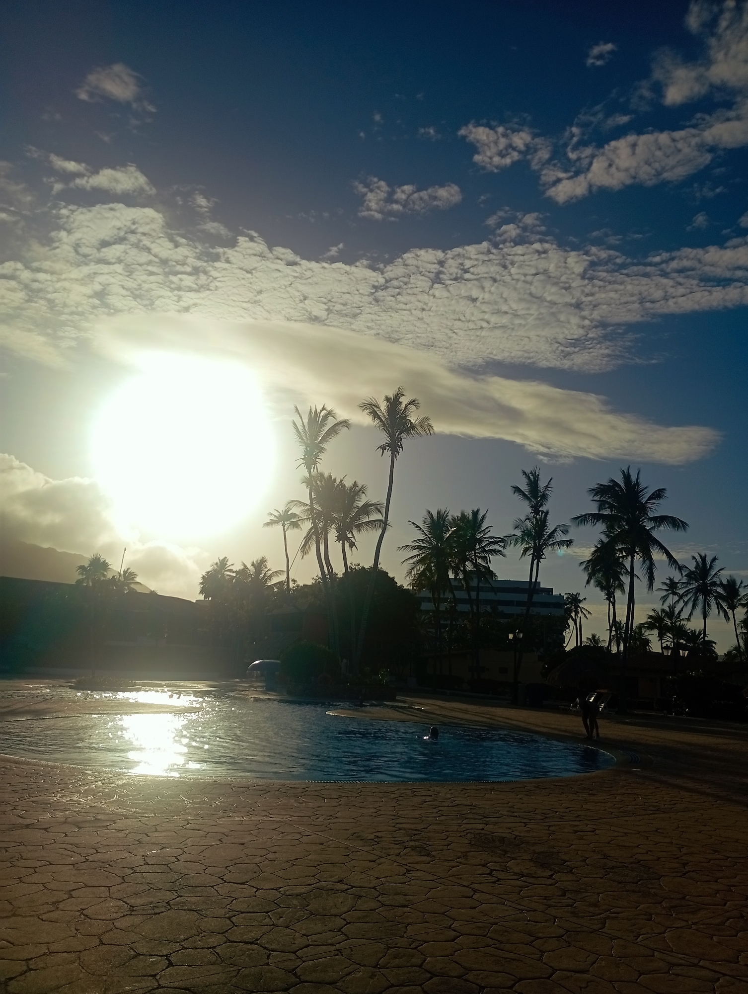 The image shows a scene at Playa El Agua, Venezuela, with a swimming pool in the foreground and palm trees silhouetted against a bright sun in a partly cloudy sky. The pool's surface reflects the sunlight, creating glare. Several people are visible in and around the pool, with one individual walking along the edge. A building is visible through the palm trees. The foreground consists of a textured, paved area.