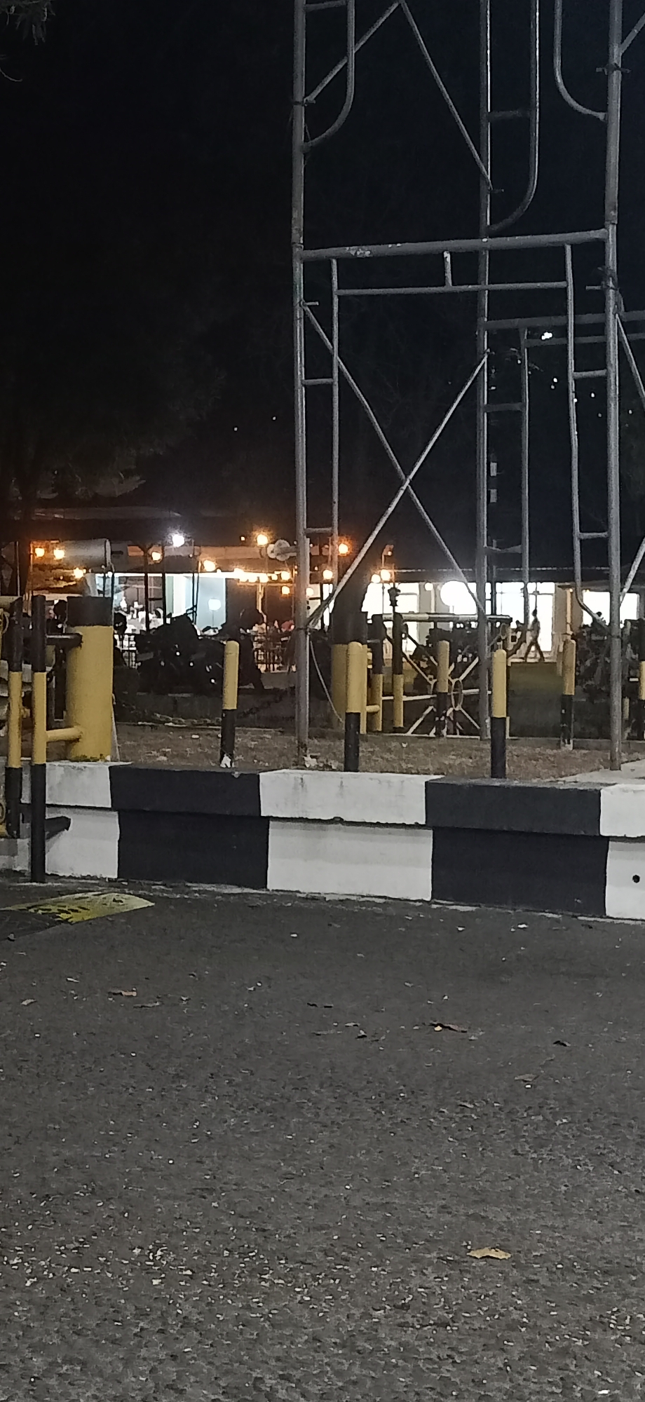 At night in Tambontunong, Indonesia, a scene unfolds near what appears to be an outdoor dining area or cafe. In the foreground, a road surface with scattered debris is visible, bordered by a concrete barrier painted with alternating black and white stripes. Behind the barrier, a dirt or gravel area contains several yellow and black bollards, and scaffolding structures made of metal pipes. Further back, through the scaffolding, a building with large windows is illuminated by numerous warm-toned lights, suggesting it is a commercial establishment. Silhouettes of people and tables are discernible within the building, implying a gathering or service activity. Trees with dark foliage are also present in the background. The overall atmosphere is dark, typical of nighttime, with artificial lighting playing a significant role in illuminating the scene.