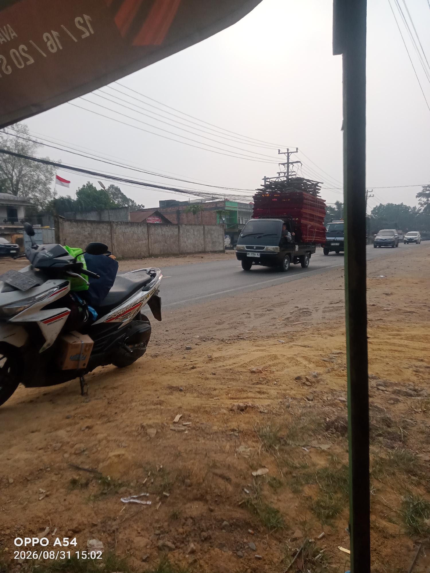 The image depicts a street scene in Jambi, Indonesia, on a hazy day. In the foreground, a white and silver scooter is parked on a dirt shoulder. A blue bag and a green bag are placed on the seat of the scooter, along with a brown cardboard box underneath the front. The road ahead has two lanes of traffic. A dark colored mini truck, heavily loaded with red and brown materials stacked high, is driving in the right lane. The truck has a license plate that appears to read "BH 9706 NJ". Behind the mini truck, two cars are visible, one dark and one lighter colored, also traveling in the same direction. In the background, there are buildings, a concrete wall, trees, and utility poles with many power lines. A red and white Indonesian flag is flying from a pole on the left side of the image. The overall environment appears to be a peri-urban or rural area with a mix of infrastructure and natural elements. The timestamp at the bottom of the image indicates it was taken on August 31, 2026, at 15:02.