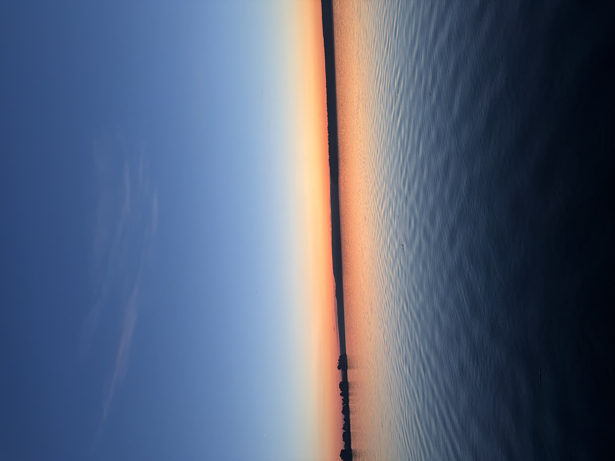 This image depicts a wide-angle view of a large body of water during either sunrise or sunset, located in Unknown, Unknown. The sky transitions horizontally from a deep indigo blue at the top, gradually lightening to a vibrant orange and red along the horizon line. Sparse, faint cirrus clouds are visible in the upper blue portion of the sky. A dark, uninterrupted silhouette of distant land, likely a treeline, delineates the horizon. The water surface mirrors the sky's coloration, displaying a gradient from a luminous orange-blue near the horizon to a dark, deep blue or black in the immediate foreground. Numerous small, parallel ripples are visible across the water's surface, particularly in the mid-ground. No human subjects, animals, or prominent man-made objects are present in the frame. The scene is illuminated by the low-angle light characteristic of dusk or dawn.