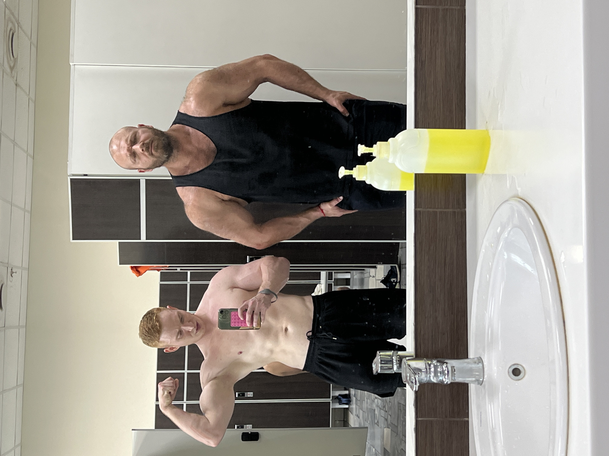 In a gym locker room, two men are reflected in a mirror. The man on the left, with red hair, is shirtless and flexing his bicep while holding a phone. He is wearing black shorts. The man on the right is wearing a black tank top and appears to be looking forward with his hands on his hips. The background shows lockers and a sink area with soap dispensers. The location is a locker room in the city of Chaikovsky, Russia.