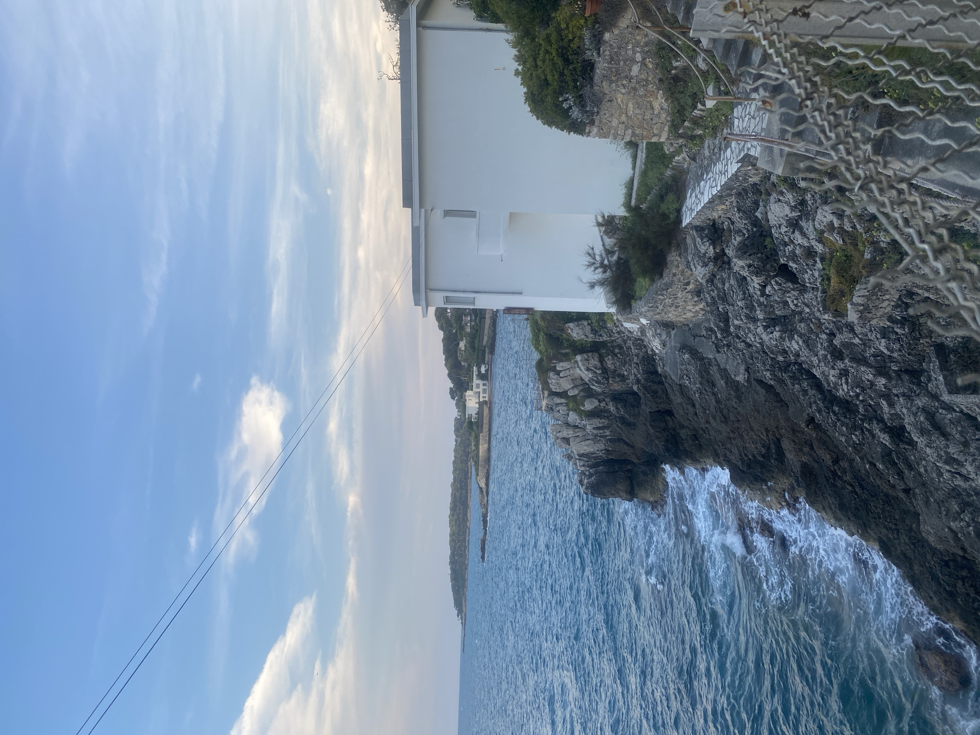 This is a photograph of a coastal scene near Antibes, France. In the foreground, a rocky cliff drops down to the sea. The water is a deep blue and appears somewhat choppy with whitecaps. A white, modern building is situated on the cliff edge, with a pathway leading down towards the sea. The building has clean lines and a flat roof. The pathway is made of stone and bordered by low stone walls and vegetation. A wire fence is visible in the lower right corner of the image. In the background, the coastline curves into the distance, with more buildings and dense green trees visible. The sky is a pale blue with scattered white clouds, and two dark lines, possibly power lines, stretch diagonally across the upper part of the frame. The overall impression is of a serene, scenic location.