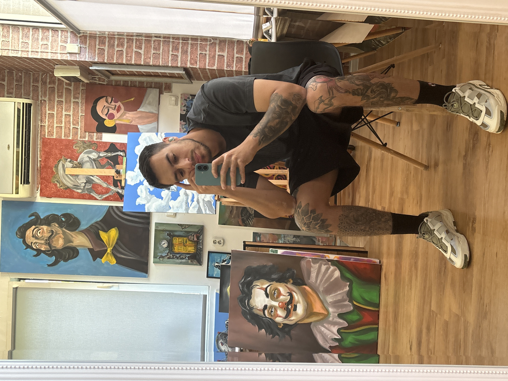 The image shows a man taking a selfie in a room that appears to be an art studio or gallery. The man is light-skinned with dark hair and a beard, wearing a dark gray T-shirt, black shorts, and black socks. He has tattoos on both arms and legs. He is holding a light blue smartphone in his left hand to take the photo.
The background reveals a brick wall on the left side of the frame, suggesting an interior setting. Numerous paintings are visible, depicting various subjects, including portraits, a horse, landscapes, and surrealist figures. A white air conditioning unit is mounted on the wall above the paintings.
The man is sitting on a wooden chair. The floor is wooden and the image is captured as a reflection in a mirror.
There's no information to confirm that the location is in the city of Plovdiv, Bulgaria, based on the visible elements in the image.