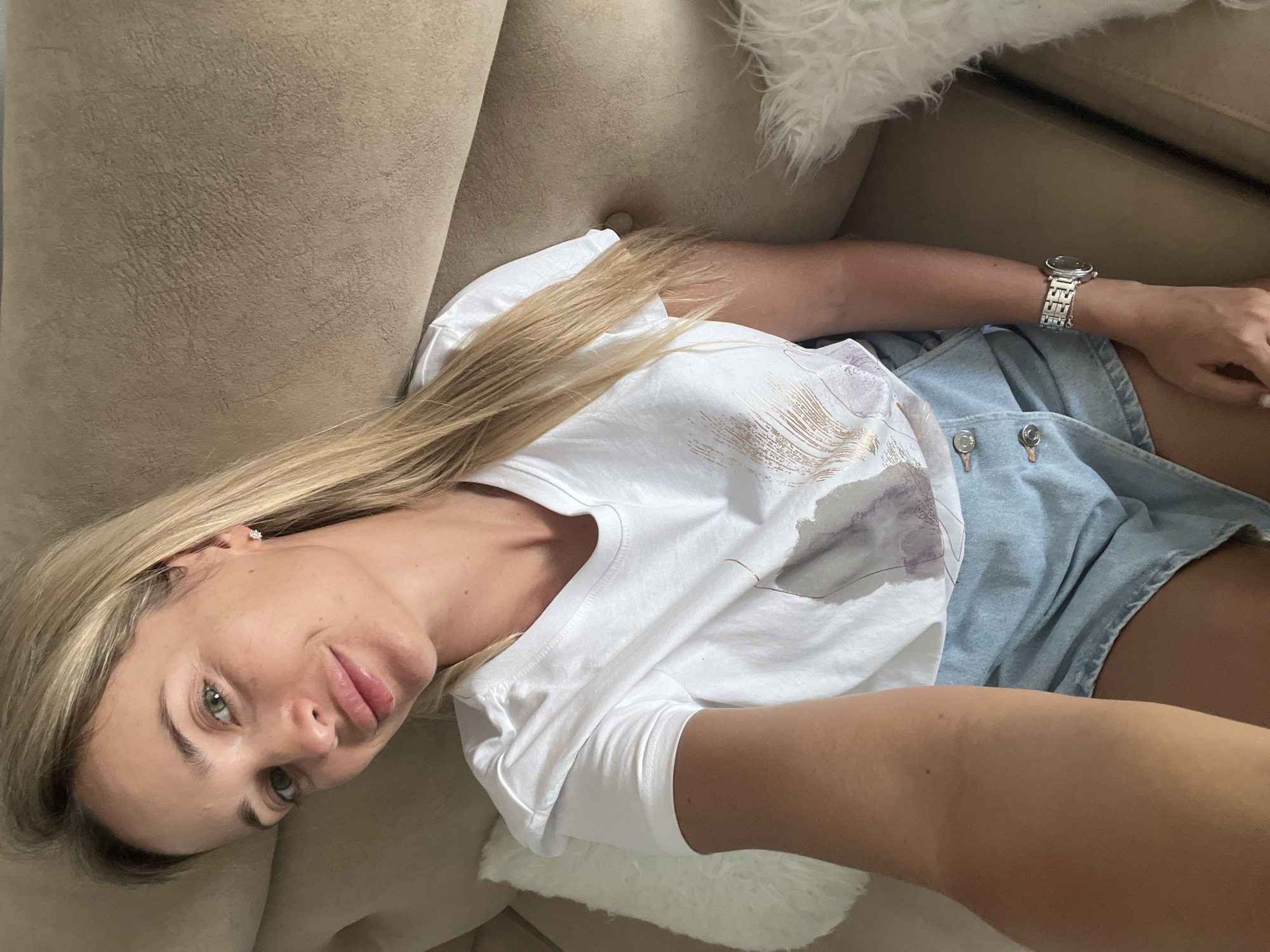 A blonde woman with light green eyes is captured in a selfie, seated on a brown sofa. She is wearing a white t-shirt with a faded gold and purple feather print, and light blue denim shorts with two visible buttons at the waist. A silver wristwatch adorns her left wrist. Two white decorative pillows are visible: one to her left, with a shaggy texture, and a portion of another to her right, also shaggy. The background consists of the textured brown sofa. The image appears to have been taken indoors, possibly in a hotel room or living area in Sharm El Sheikh, Egypt.