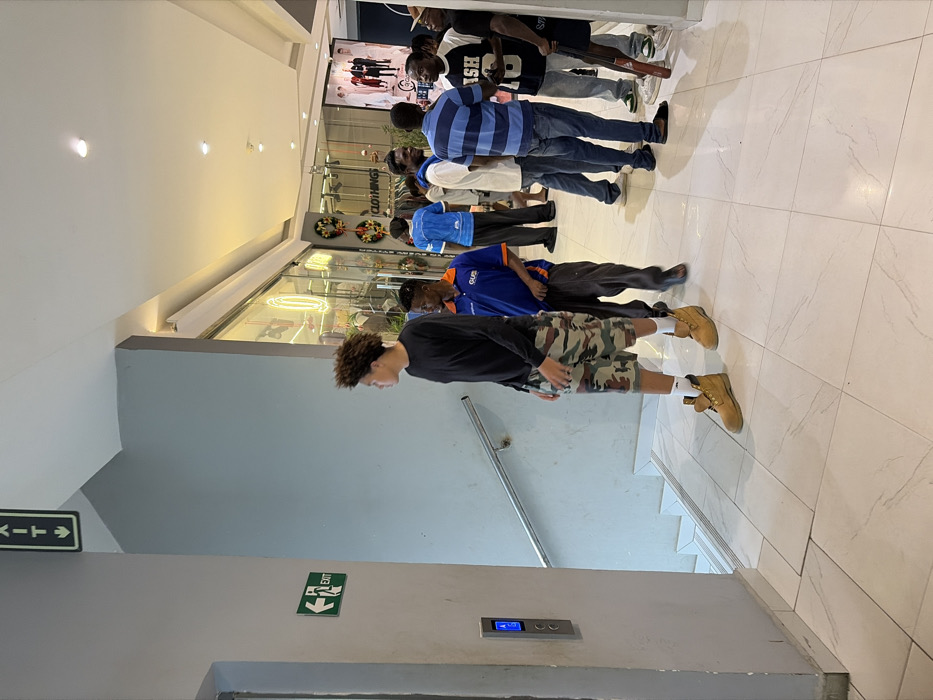 A group of people stand in line within a building. An individual wearing a black shirt, camouflage shorts, and tan boots is positioned near an elevator and stairs. An exit sign is visible on the wall. The remaining individuals are in front of a store with clothing displays. One person holds a baseball bat. The floor is tiled, and the building's interior appears to be modern with good lighting.