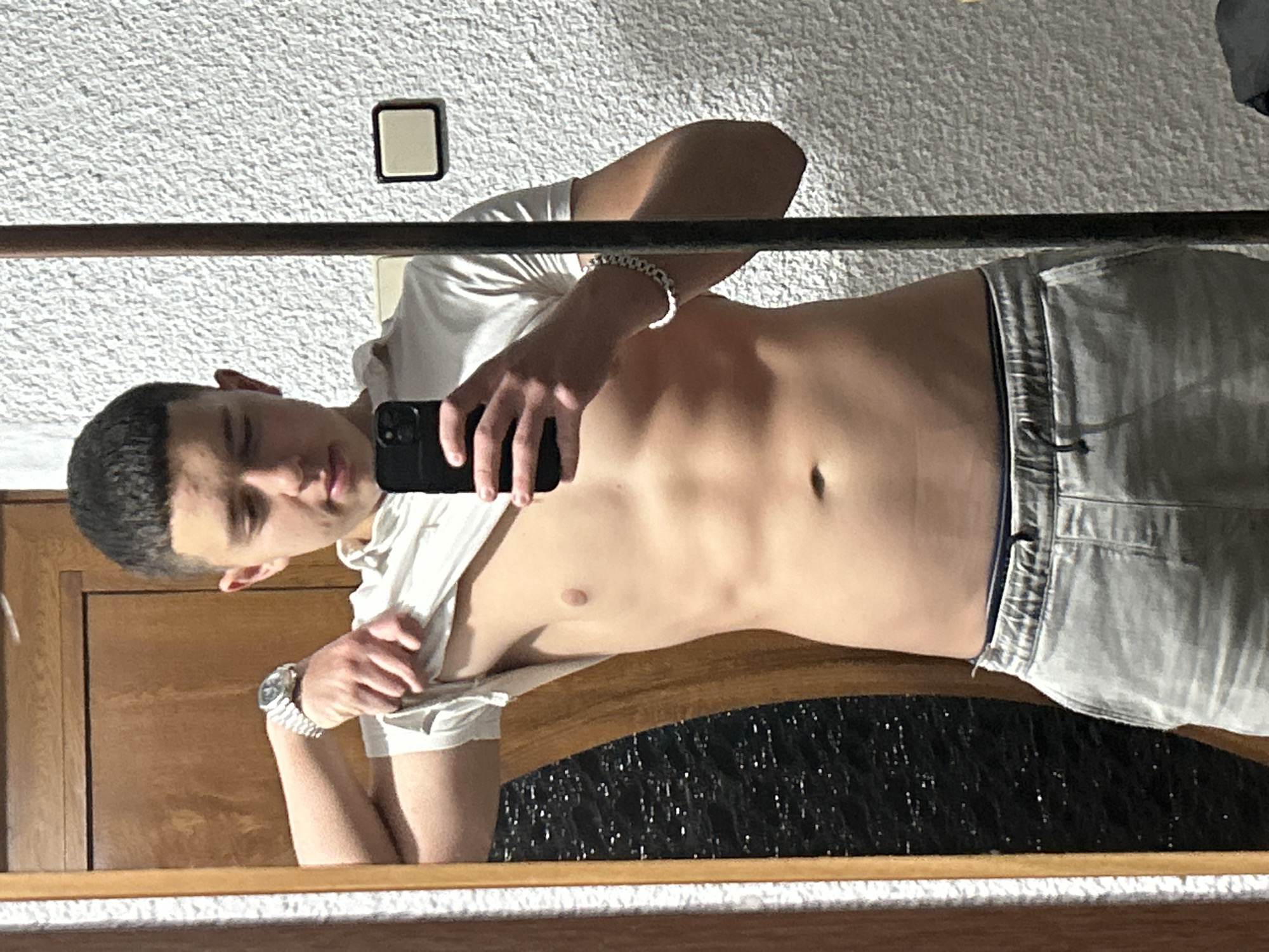 A male, reflected in a mirror, is holding a black smartphone in his right hand and partly lifting the front of his white t-shirt with his left hand. He is wearing light-colored shorts with a dark waistband and a silver bracelet on his right wrist. He also appears to be wearing a watch on his left wrist. The background is textured, with a light switch on the wall. The mirror frame is visible, and there is what appears to be a wooden door or cabinet behind him and below. The location cannot be ascertained as being in Zlatograd, Bulgaria with the available image content.