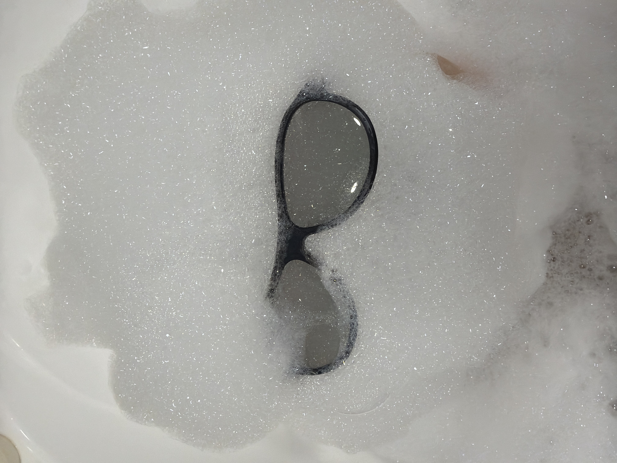 The image depicts a pair of black-framed sunglasses submerged in a white, foamy substance. The foam appears to be bubbles, possibly from soap or a cleaning agent. The background and surrounding area consist of a white, smooth surface, likely a bathtub or sink. There are no people visible. The location cannot be determined to be Votkinsk, Russia, or any other specific place from the given image.