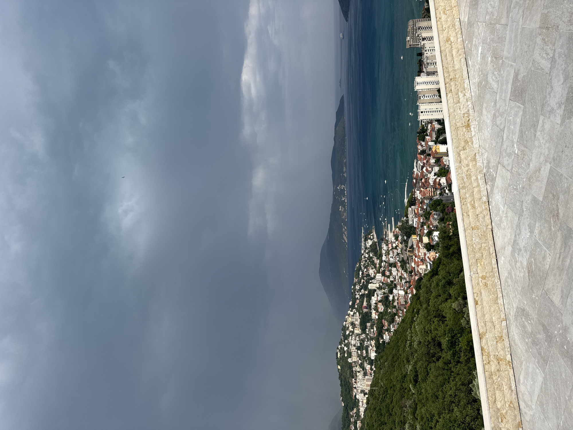 This media file shows a scenic view of Igalo, Montenegro. The scene is dominated by the sparkling blue sea on the right, meeting a coastline dotted with buildings and lush green hills. The architecture appears to be a mix of modern, white buildings and older, red-roofed houses, suggesting a vibrant town nestled along the water.
In the foreground, a textured stone pathway creates a boundary, leading the viewer's eye towards the expansive vista. The sea is calm, with several boats visible, indicating a leisurely day. The weather is dynamic, with a dramatic contrast between the bright sky and a dark, stormy cloud formation encroaching from the left. This juxtaposition of light and dark weather adds a sense of depth and drama to the scene.
The time of day is difficult to determine precisely due to the varied lighting conditions, but the presence of both bright sunlight and approaching storm clouds suggests it could be late afternoon. The overall mood is one of natural beauty and the power of the elements, with a hint of impending weather change. There is no visible text in the image.