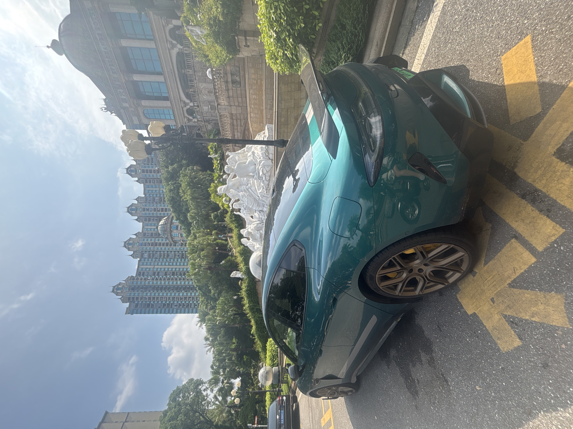 A side-rear view of a green electric sedan is captured in the city of Nanhai District, China. The car is parked on a street with yellow directional markings painted on the asphalt. In the background, several tall buildings with ornate architecture are visible, along with lush green trees and a large fountain featuring white sculptures. The sky is partly cloudy and bright, indicating a daytime scene. A black sedan is parked further down the street. The overall environment suggests a well-maintained urban area with a mix of modern and classical architectural styles.