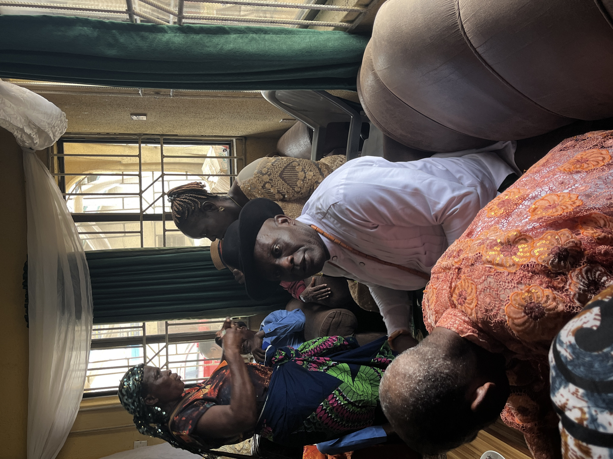 A low-angle medium shot shows a group of people in Warri, Nigeria. A man in a white shirt and black hat faces the camera with a slightly tilted head. A woman in a patterned dress and headscarf lies in a seated position. The rest of the people are obscured by the foreground. A window with bars is visible in the background. There is a dark green valance at the top of the window. A sofa is visible.