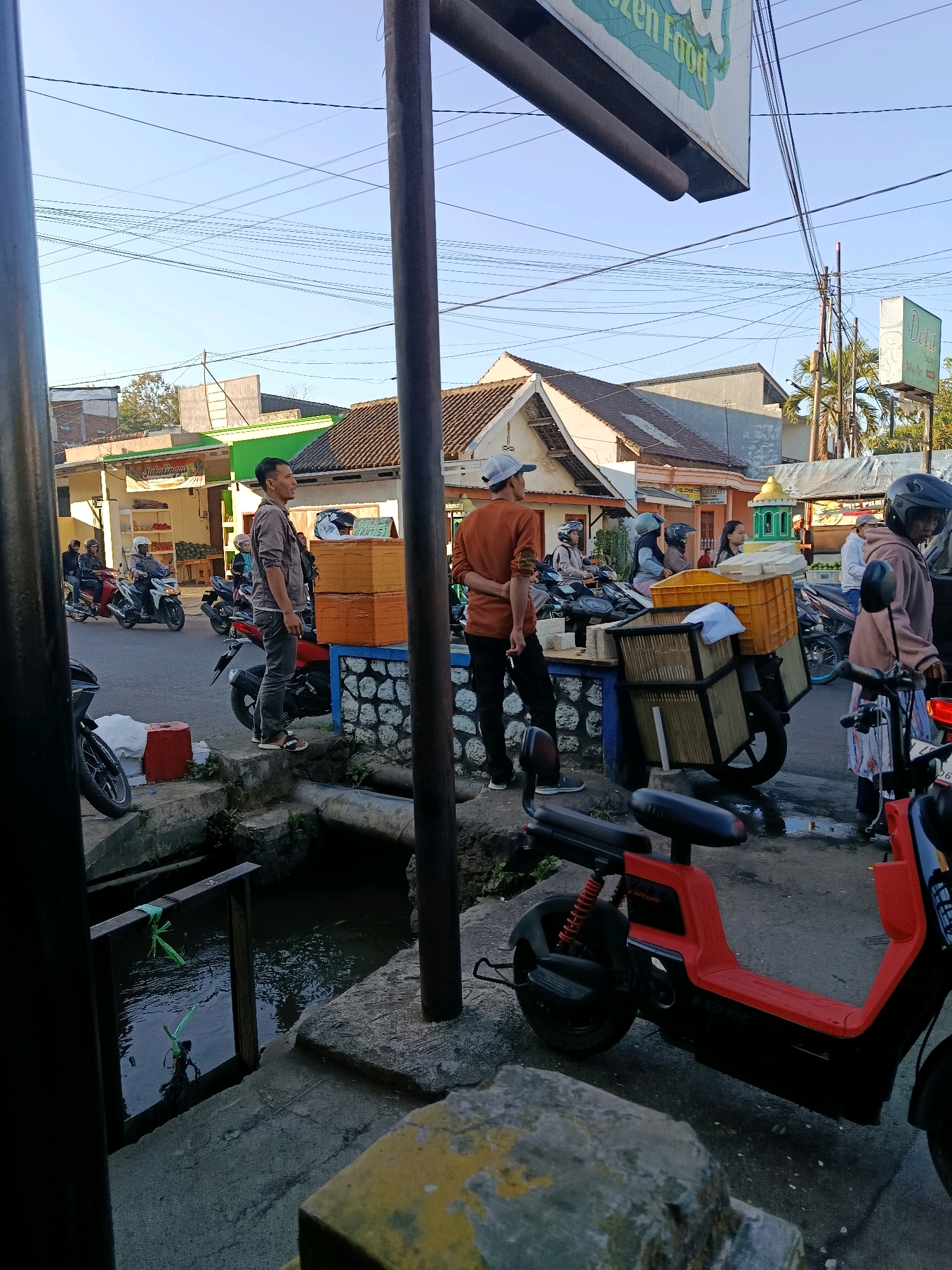 This image captures a street scene in Bululawang, Indonesia, on a sunny day with clear skies. In the foreground, a red electric scooter is parked. To the left, a small, murky canal runs alongside a concrete walkway. A wooden railing with green rope is visible over the canal. A person stands on the walkway, looking to their left. Further back, a street is lined with buildings and shops. Several motorcycles and scooters are on the road, with some people riding them and others standing nearby. One person, wearing an orange shirt, is standing on the walkway looking away from the camera. Another person, wearing a grey jacket, is also standing on the walkway. A motorcycle with stacked orange boxes is parked on the side of the road, along with another motorcycle carrying a yellow crate. Signs are visible on the buildings, one reading "Frozen Food" and another with the word "Deta". The overall scene depicts a typical urban environment with pedestrian and vehicular traffic.