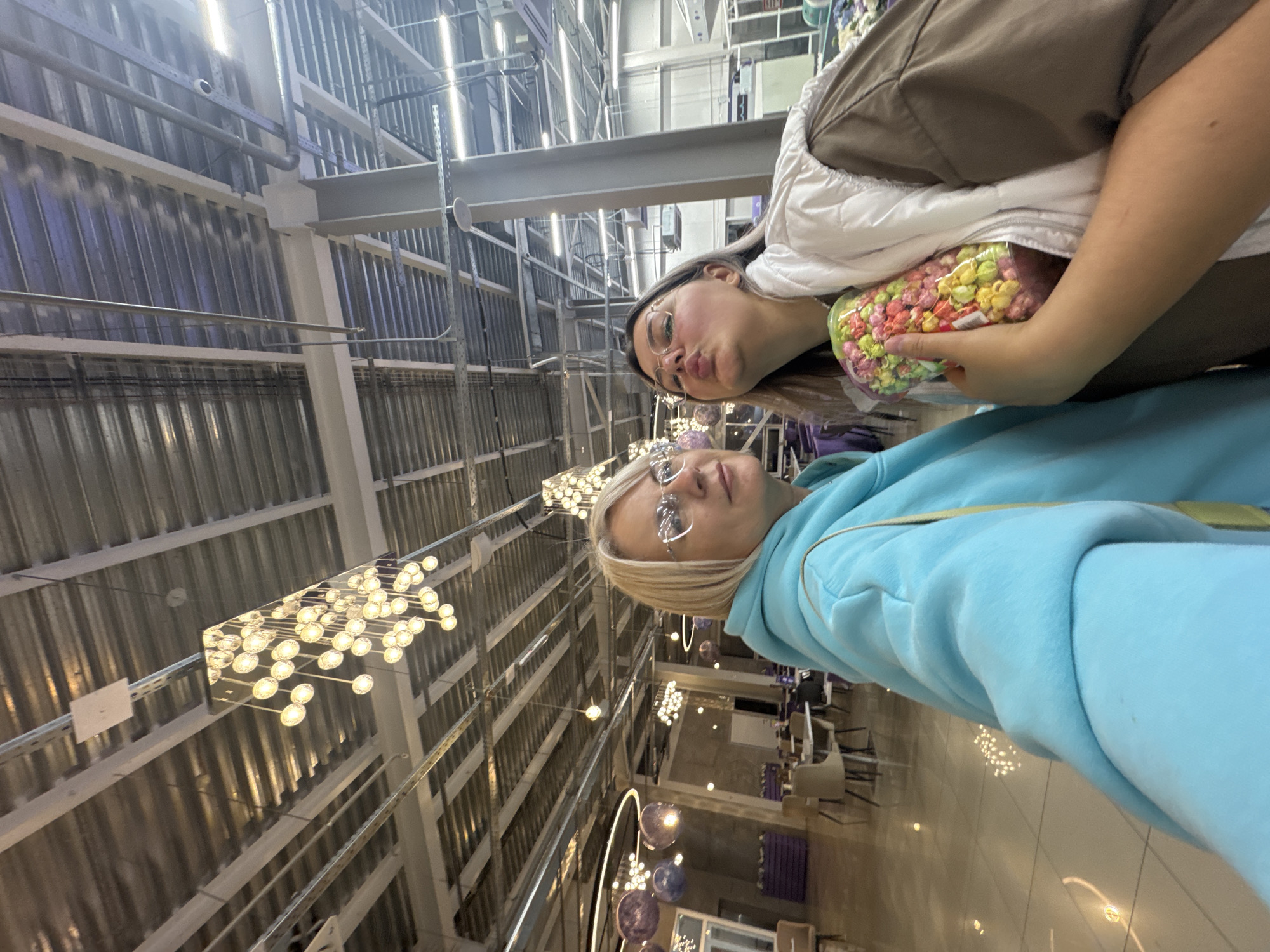Two women pose in an indoor space in what appears to be a store or commercial building in Birobidzhan, Russia. The first woman, wearing glasses, is in the foreground, wearing a light blue hoodie. The second woman behind her is holding a clear container filled with colorful popcorn or candy. She wears a white quilted jacket over a brown shirt, and also has glasses. The background reveals an industrial-style ceiling with metal beams, visible ventilation, and unique light fixtures. There are also rows of tables and chairs suggesting a dining area or cafeteria.