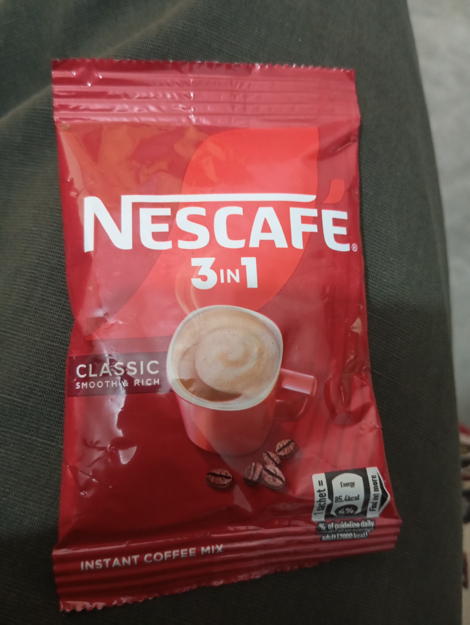 The image shows a close-up of a red Nescafé 3-in-1 Classic instant coffee mix sachet. The sachet is placed on a dark, textured fabric, possibly clothing. The Nescafé logo is prominently displayed in white, with "3-IN-1" below it. A graphic of a red cup filled with coffee is on the right side, along with coffee beans. Text on the lower left reads "CLASSIC SMOOTH & RICH" and on the bottom edge, "INSTANT COFFEE MIX". On the bottom right, there is a nutritional information label indicating "Energy 85.4 kcal" and "% of guideline daily adult (2000 kcal)". The location is described as Unknown, Unknown.