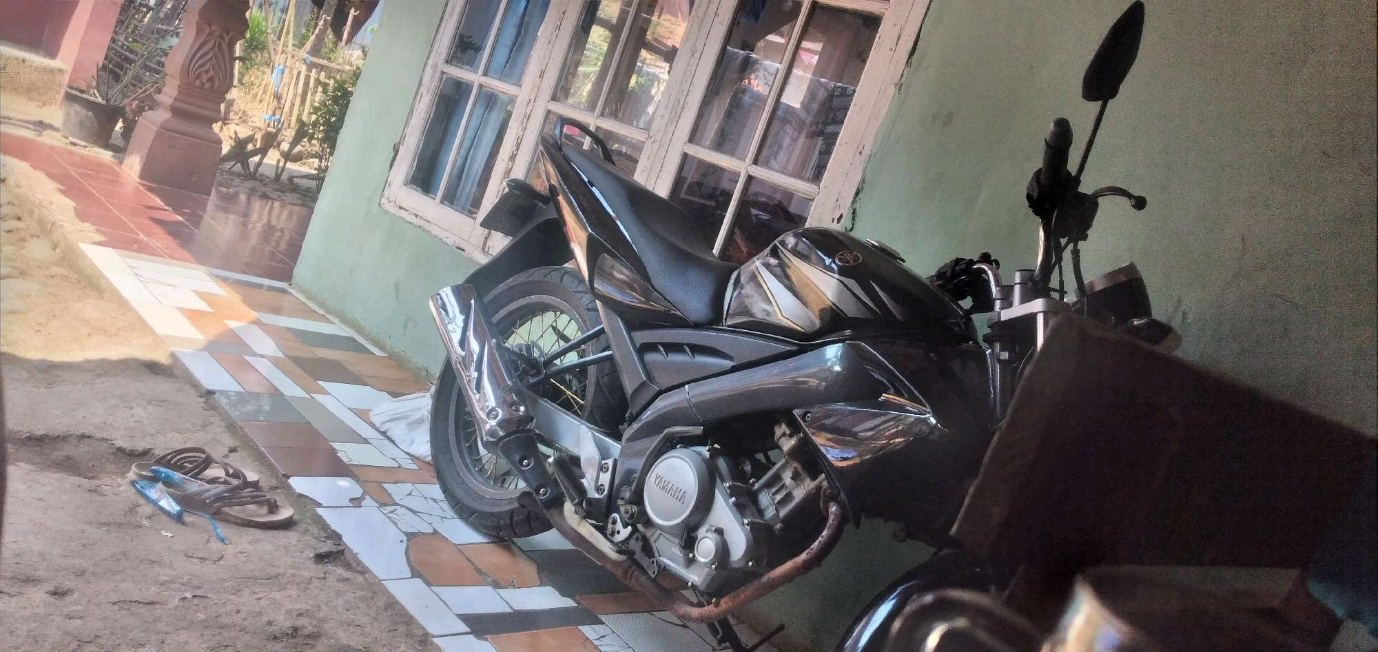 In Rahong, Indonesia, a black Yamaha motorcycle is parked on a tiled pathway adjacent to a light green wall. The motorcycle is angled, with its rear wheel and exhaust pipe prominently visible. The engine casing displays the "YAMAHA" logo. To the left of the motorcycle, a pair of brown sandals lies on a dirt ground. Behind the motorcycle, a window with multiple panes is set into the light green wall. The pathway is laid with rectangular tiles in various earth tones. A decorative red tiled walkway leads away from the scene, bordered by a concrete pillar. In the background, vegetation and wooden fencing are visible under bright sunlight.