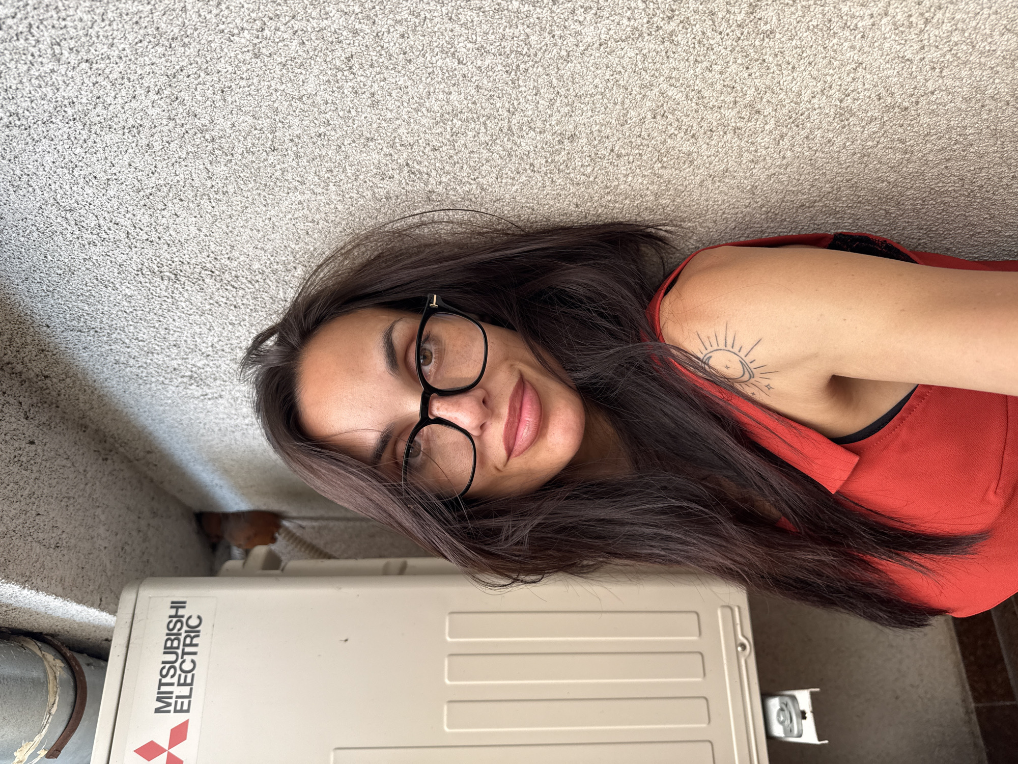 A brunette woman with glasses is posing for a selfie next to a Mitsubishi Electric air conditioning unit, presumably outdoors. The location is likely Pleven, Bulgaria, although this is not explicitly confirmed in the image. The woman is wearing a red top and has a tattoo on her shoulder. The background shows a textured wall.