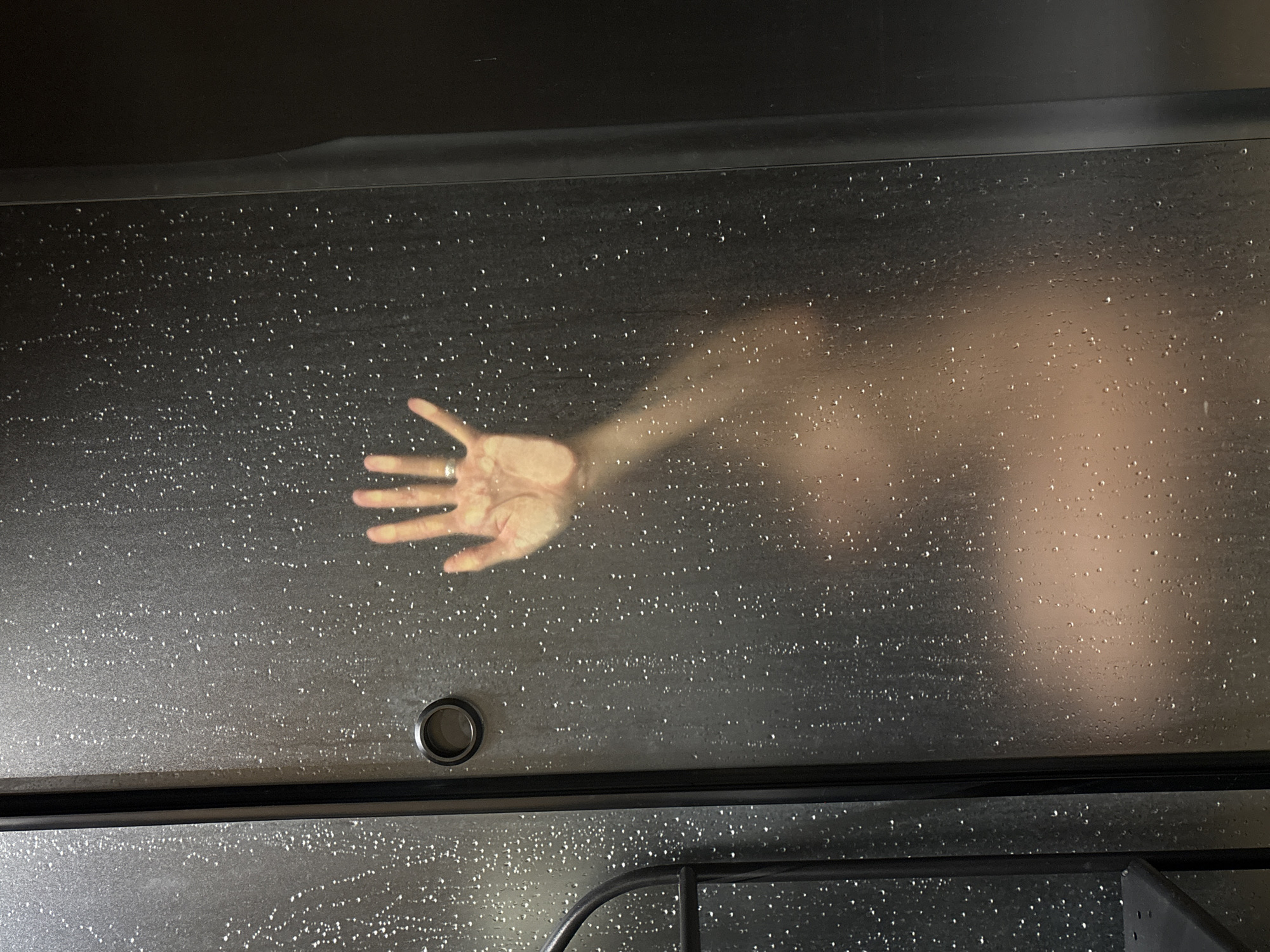 The image captures a human hand and the blurred outline of a torso pressed against a translucent, wet surface, likely a shower door or sauna panel. The surface is dark grey or black and heavily speckled with numerous small water droplets, creating a shimmering effect. The hand, appearing from the left side of the frame, is a left hand with the palm facing the viewer, and a ring is visible on the fourth finger. To the right of the hand, a diffused, lighter-toned shape suggests the presence of a person's body behind the wet screen.
Below the main wet panel, a thick, dark horizontal bar separates it from another section of a similar wet, speckled surface. In the lower right corner, part of a dark, curved metal structure is visible, possibly a handle or support. To the left of the hand on the main panel, there is a circular, dark metallic fixture, which might be a knob, a drain cover, or a connection point.
The scene depicts an individual inside a steamy or wet enclosure, interacting with the barrier. The overall lighting is dim, emphasizing the contrast between the glowing hand and the dark, wet surface. The presence of water droplets suggests a humid environment, such as a shower or steam room. There is no visible text in the image. The location context provided is Chaikovsky, Russia, though the image itself contains no specific geographical markers.
