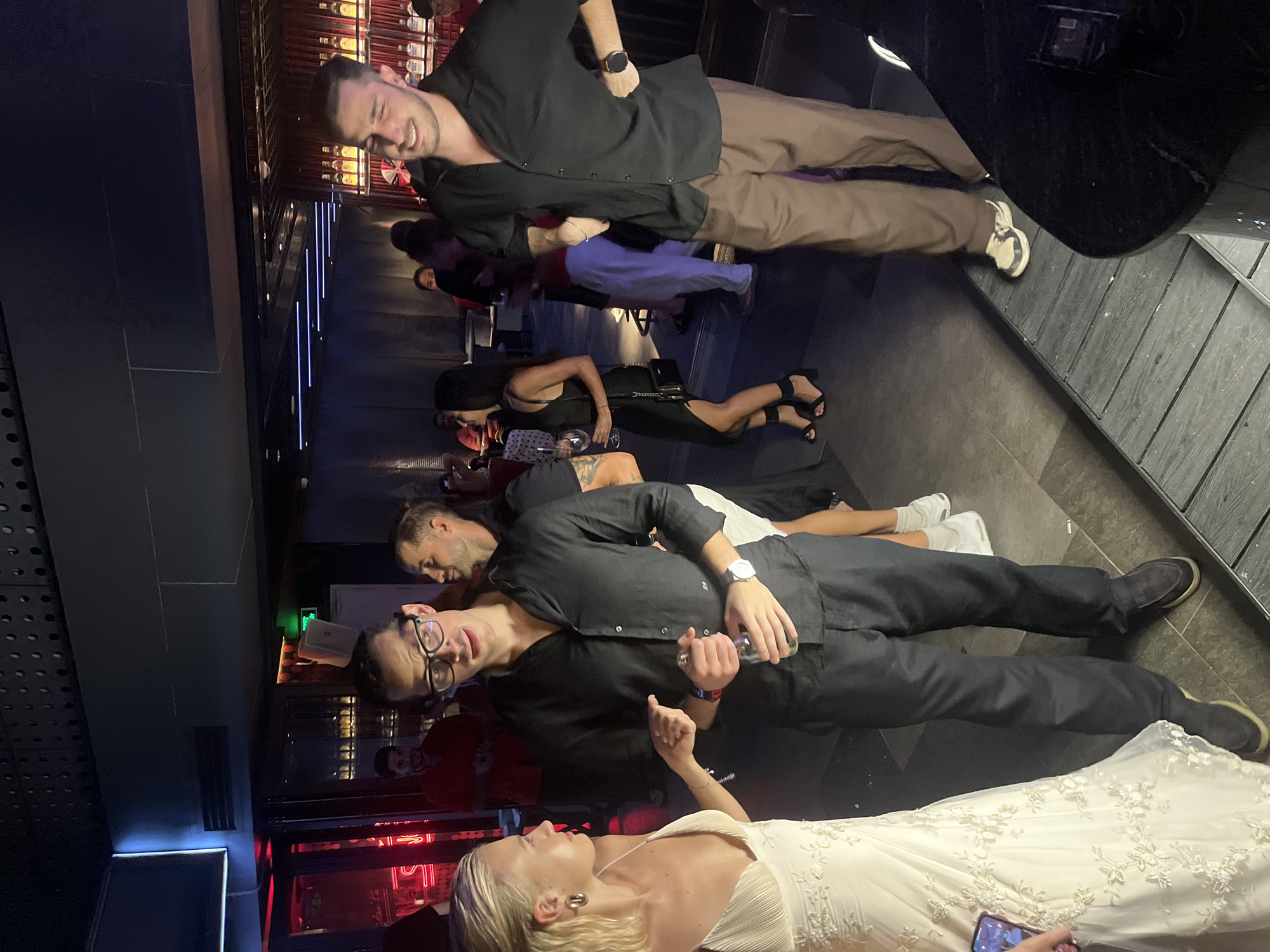 The image depicts an indoor scene, possibly a bar or club, with a group of people socializing. The lighting is dim and atmospheric, with some red and blue neon accents visible.
In the foreground, a woman in a white, ornate dress is speaking with a man wearing a black button-down shirt and black pants. The woman is holding a phone in her left hand. The man is holding a clear bottle in his right hand and appears to be listening. Both individuals are facing each other.
To the right of this pair, another man in a black button-down shirt and brown pants stands with his hand on his hip, smiling and looking towards the right side of the frame.
In the background, several other individuals are visible, some standing and some sitting. The environment suggests a social gathering. The presence of a bar area with bottles on display is implied by the shelves visible behind the standing individuals.
There is no information within the image to suggest the location is in the city of Đà Nẵng, Vietnam.