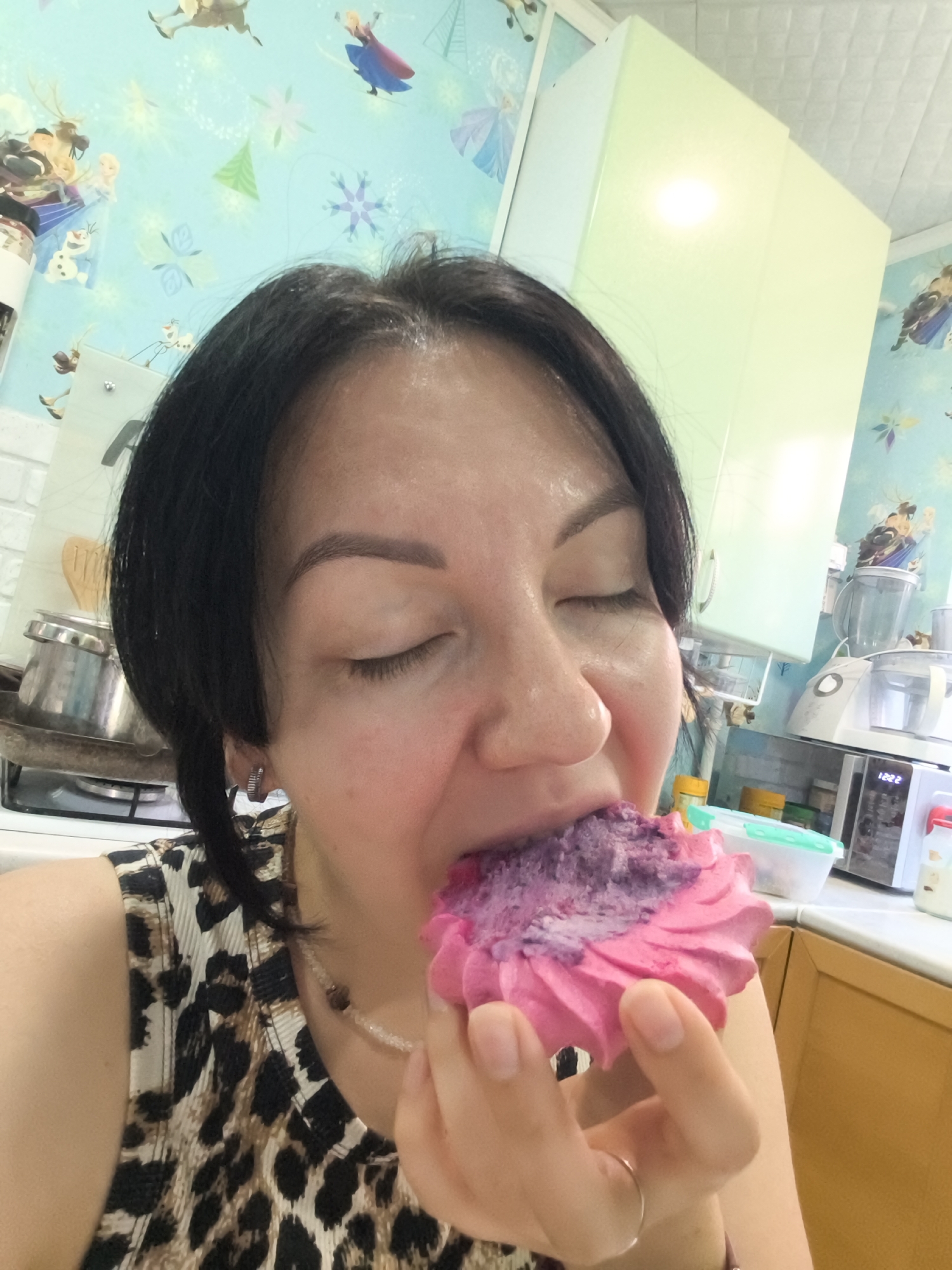 A woman is eating a pink and purple confection in a kitchen. The woman has dark hair, is wearing a leopard-print top, and a beaded necklace. The kitchen has light green cabinets, a light blue tiled backsplash with a Frozen theme, and a microwave. The time on the microwave reads 12:22. The image is taken from a medium close-up perspective. The location is not specified in the image.