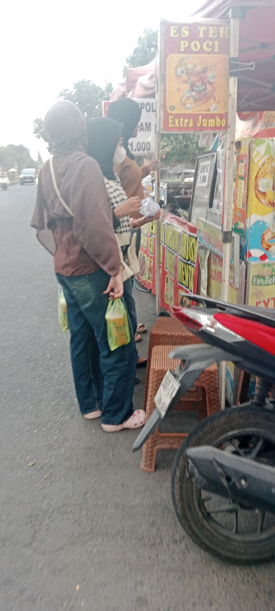 The image captures a street scene in Ngemplak, Indonesia, likely near a food stall. In the foreground, the rear wheel and fender of a motorcycle are visible, resting on a brown woven plastic stool. To the left, a person wearing blue jeans and pink clogs stands with two plastic bags of drinks in their hands. Behind them, two other individuals are visible, both wearing headscarves. One person is wearing a brown tunic and the other a checkered sweater. They appear to be interacting with a food stall. The stall has colorful banners advertising "ES TEH POCI" and "Extra Jumbo," along with prices like "1.000." In the background, trees and a road with vehicles can be seen, indicating an outdoor urban environment. The lighting suggests it is daytime.