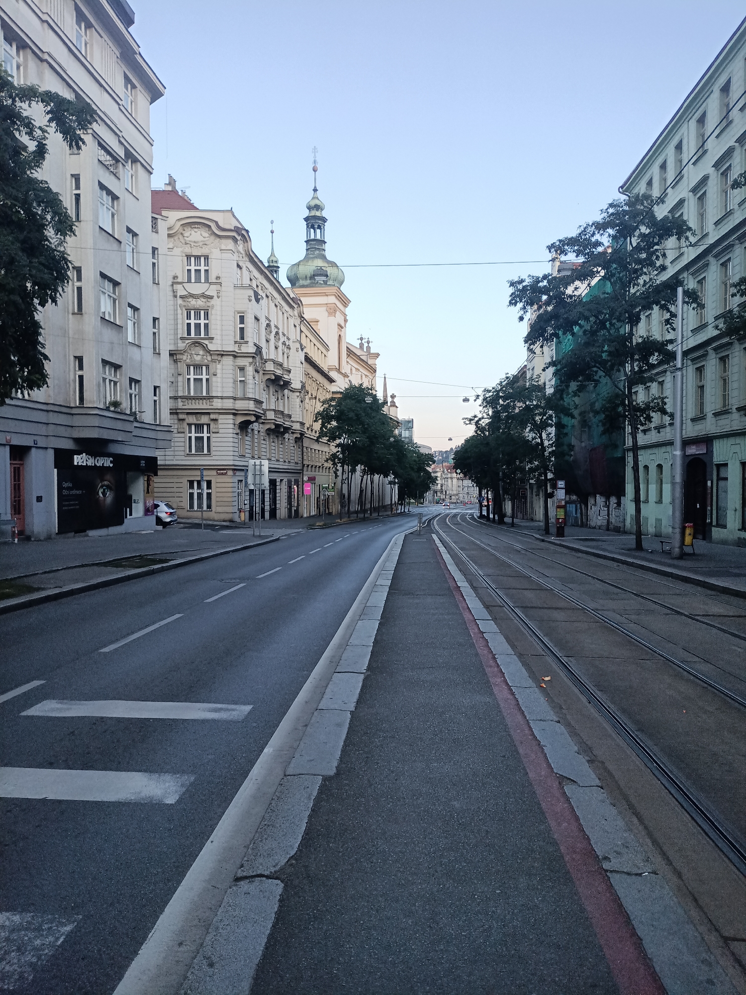 The image shows a wide street in Prague, Czechia, with tram tracks running down the middle. The street is lined with multi-story buildings on both sides, displaying architectural styles typical of European cities. On the left side, a building with a large advertisement for "PRISM OPTIC" is visible, with a white car parked in front. Further down the street, pedestrian crossings are marked with white lines. The right side of the street features more buildings, some with visible shopfronts. Trees are planted along the sidewalks. The sky is clear and a pale blue, suggesting it is either early morning or late evening. The street appears to be relatively empty of people and traffic, giving a sense of quietness. The tram tracks are prominent, indicating that trams are a common mode of transportation in this area. The overall scene depicts a typical urban streetscape in Prague.