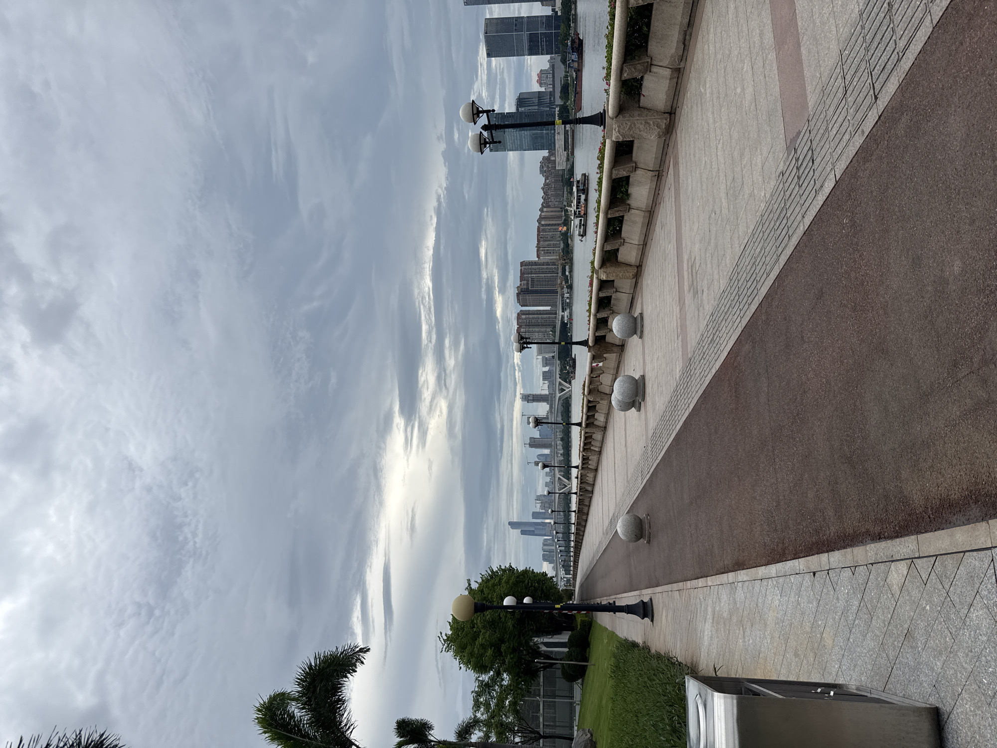 This is a vertical, eye-level, full shot of a waterfront promenade in Tianhe District, China, under an overcast sky. The promenade, paved with light gray and red-brown sections, features a walking path and a bikeway. Decorative spherical bollards are spaced along the path. On the left, a lush green lawn borders the promenade, with a metal trash receptacle visible near the bottom left corner. Palm trees are present on the far left.
Across the water, a city skyline is visible, characterized by numerous tall buildings, some under construction, and a bridge. Several boats, including a white passenger boat and a dark cargo vessel, are on the water. Lampposts with multiple globes are positioned along the promenade, and the waterfront is lined with a stone retaining wall. The overall atmosphere is serene, with no people visible in the frame.