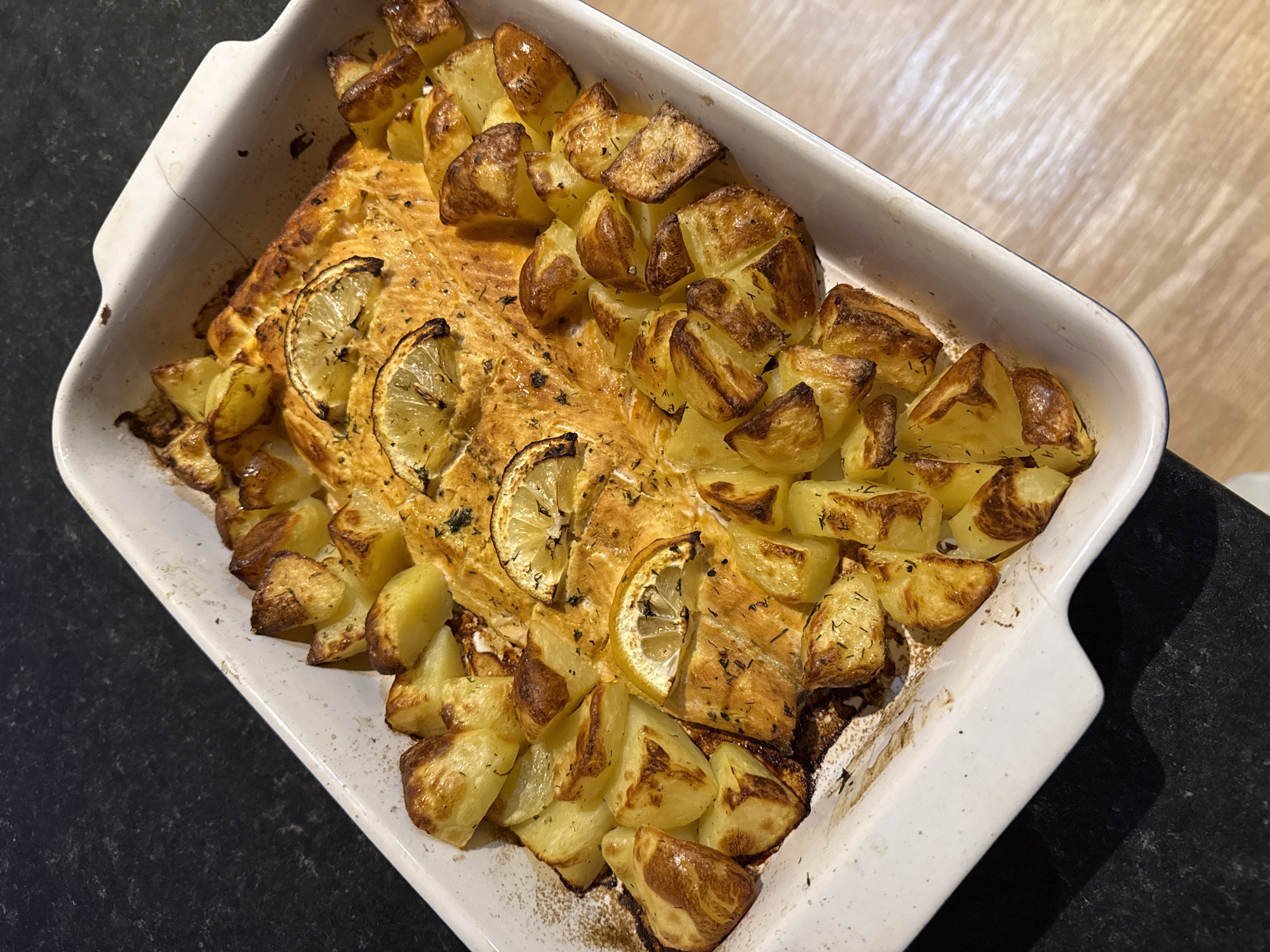 The image shows a white rectangular baking dish filled with roasted food. A piece of fish, possibly salmon, is in the dish, garnished with lemon slices. Roasted potatoes are scattered around the fish. The baking dish is sitting on a surface that appears to be part of a counter or table, with a wooden-like grain visible on one side and a darker surface on the other. The scene suggests a home-cooked meal. There is no indication of the location being Great Yarmouth, United Kingdom, or any specific city.