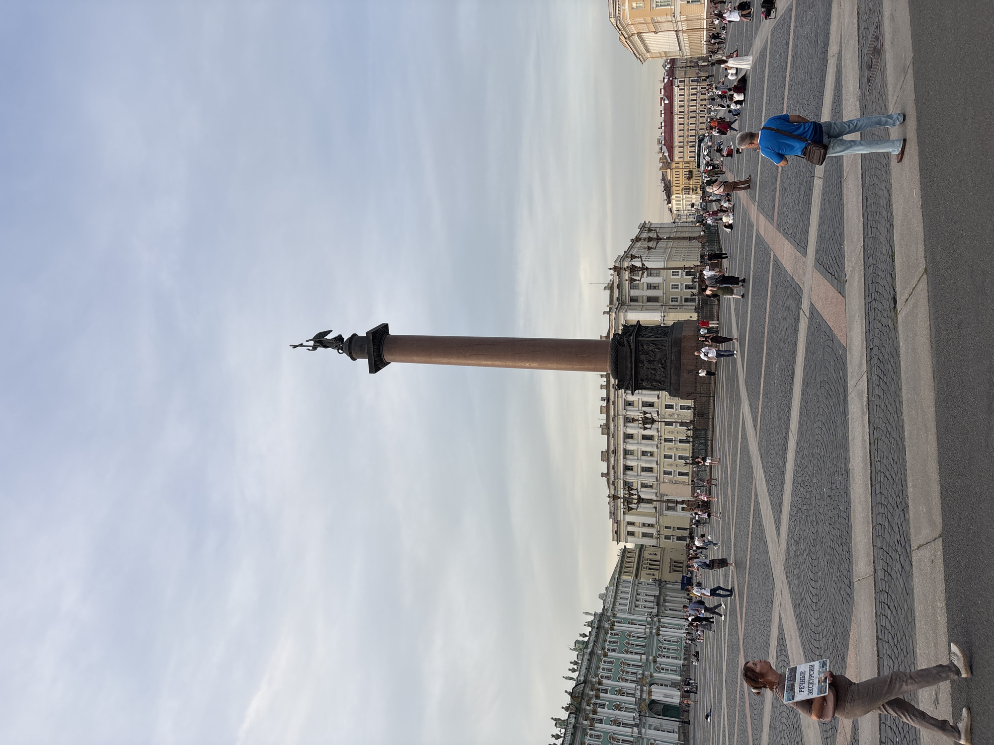 The image depicts a wide, paved public square in Saint Petersburg, Russia, likely Palace Square, given the presence of the Alexander Column. The sky is overcast with diffused light. Dominating the center of the square is the tall, reddish-brown Alexander Column, topped with a statue of an angel. In the background, the ornate buildings of the General Staff Building and the Winter Palace are visible.
Numerous people are scattered throughout the square, engaged in various activities. Some are walking, while others are sitting or standing in groups. In the foreground, a woman walks from left to right, carrying a sign with Russian text that reads "Речные Экскурсии" (River Excursions). To the right, a man in a blue shirt and jeans stands looking towards the Alexander Column. The paving stones of the square are laid in a patterned design. The overall scene is that of a busy tourist location with a mix of individuals and small groups enjoying the open space.