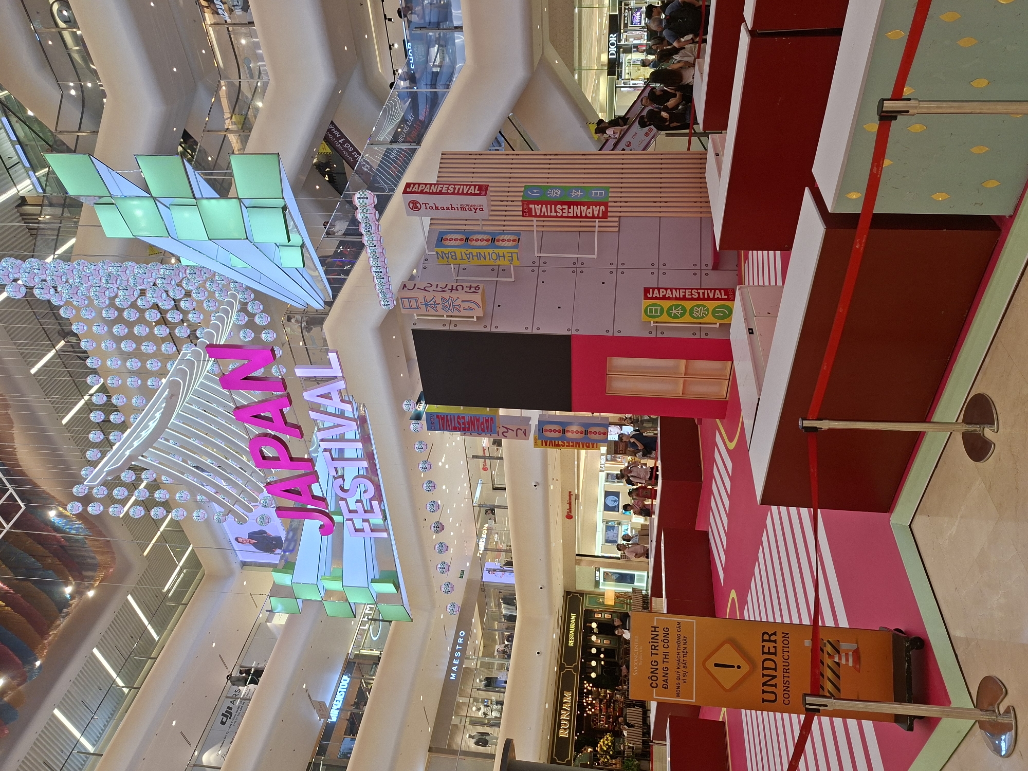 The image depicts the interior of a shopping mall in Thủ Đức City, Vietnam, specifically at Saigon Centre. A large "JAPAN FESTIVAL" sign is prominently displayed overhead, illuminated in bright pink and white neon. The festival setup includes a decorative, multi-tiered structure resembling a stylized building with Japanese characters and colorful banners. Various shops are visible on different levels of the mall, including "RUNAM RESTAURANT", "MAESTRO", "Takashimaya", and "DIOR". In the foreground, a section of the mall is cordoned off with red tape and stanchions, with an "UNDER CONSTRUCTION" sign in Vietnamese and English. Several people are scattered throughout the mall, some browsing in shops, others gathered near the festival display, and some walking on escalators. The overall environment is brightly lit, suggesting daytime or well-lit interior lighting.