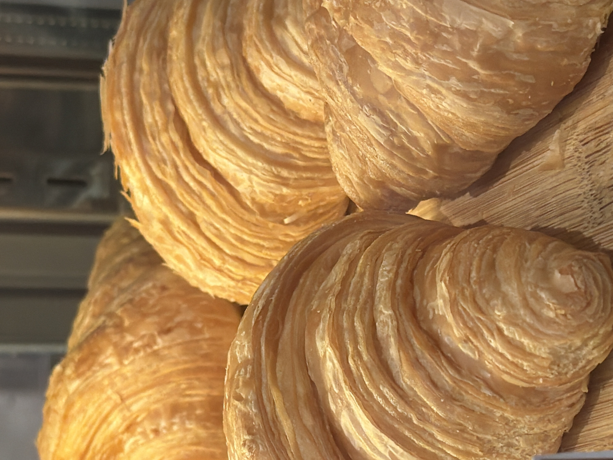 A close-up, eye-level shot captures a quartet of golden-brown croissants arranged on a wooden surface. The croissants, each with a visibly flaky, layered texture, are slightly overlapped, creating a sense of abundance. The lighting highlights the crisp edges and the warm, inviting hue of the pastries. The background is blurred and dark, suggesting an indoor setting such as a bakery or cafe. This image was taken at a cafe in Nha Trang, Vietnam.